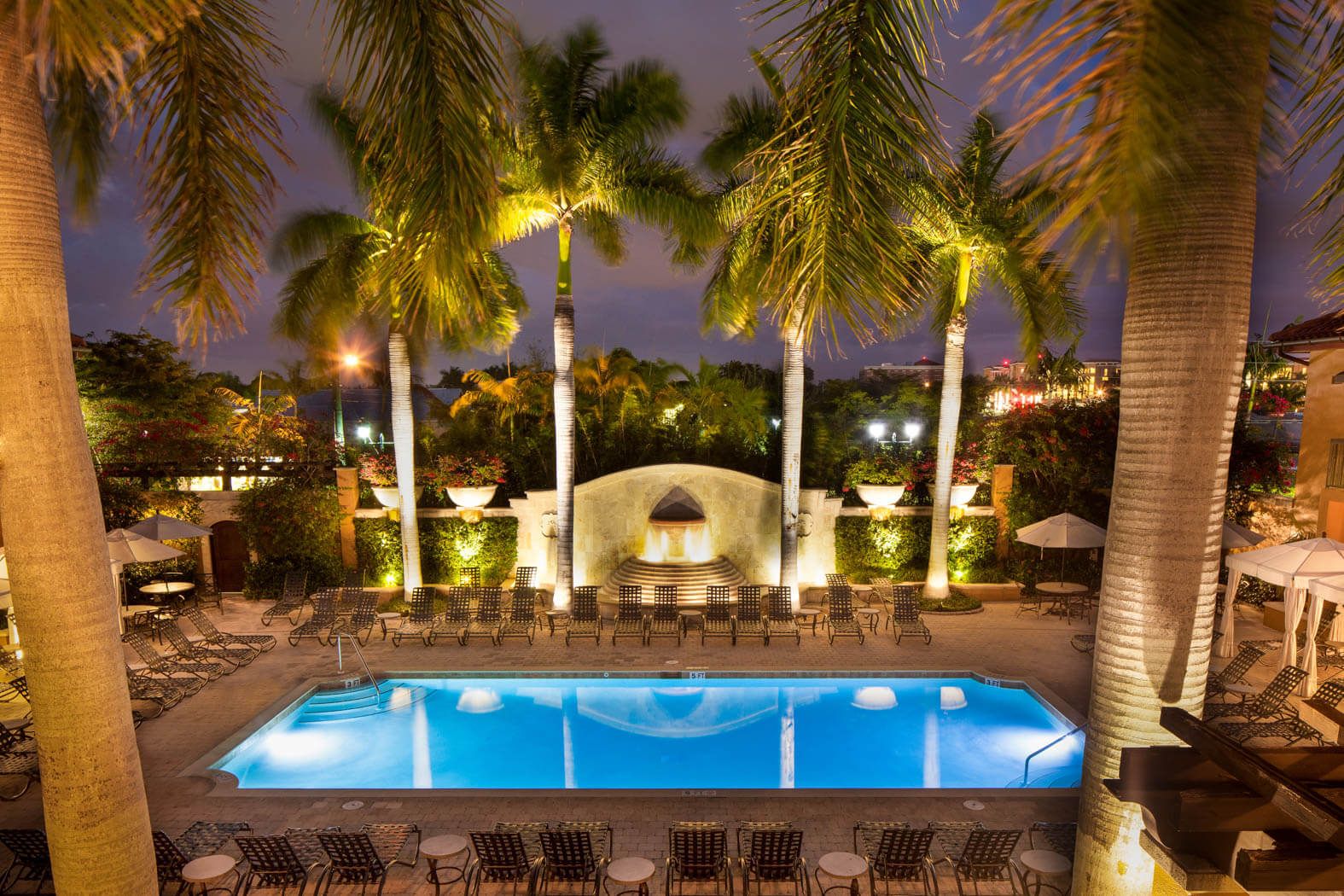 Nighttime pool courtyard with palm trees, lit lounge chairs, and a glowing blue swimming pool