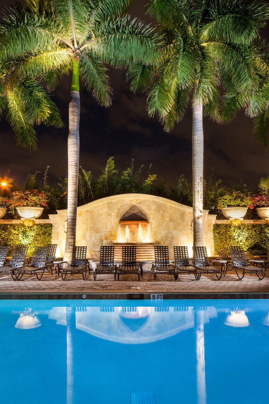 Nighttime poolside patio with palm trees, lounge chairs, and a lit stone backdrop reflected in blue water
