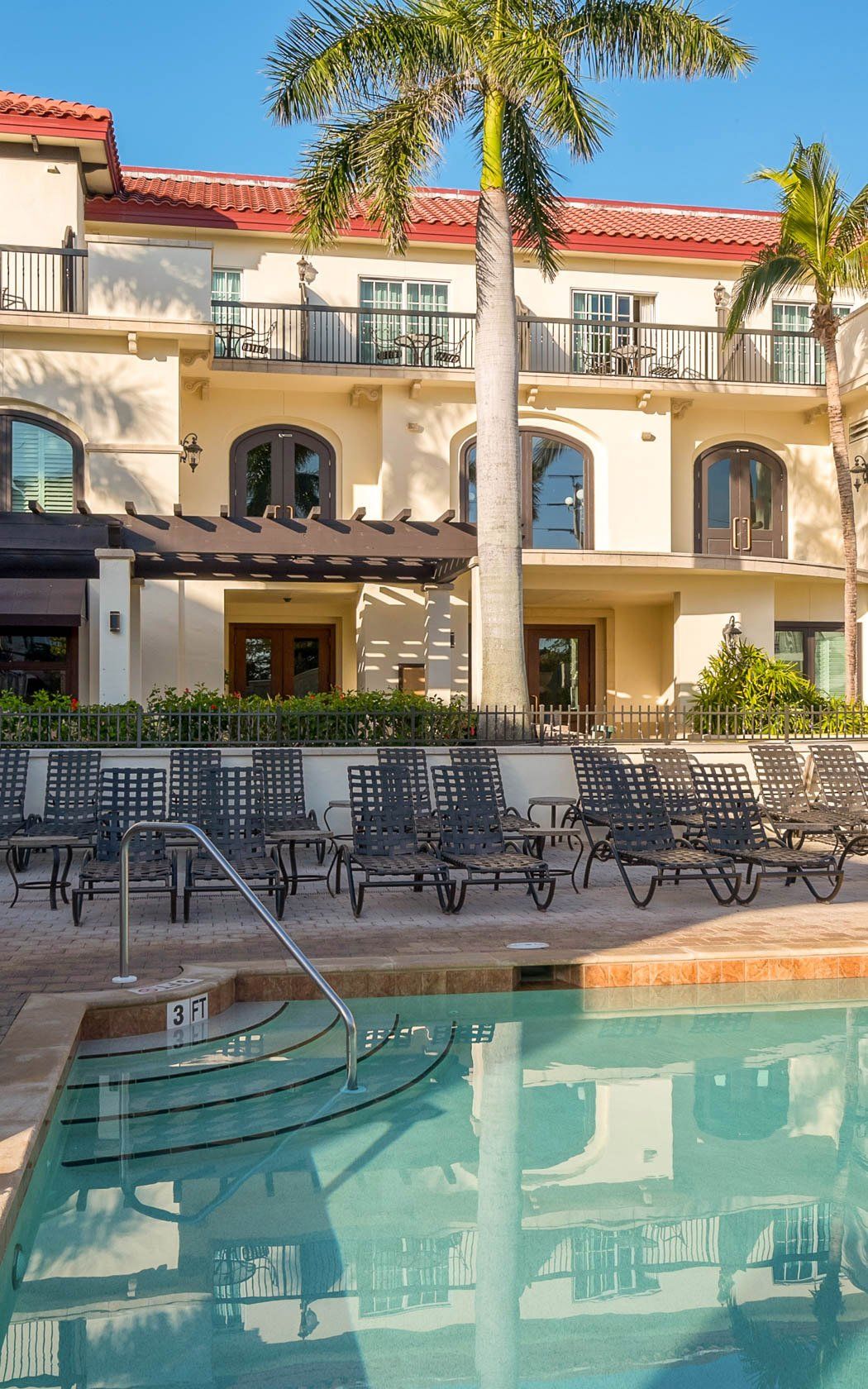 Sunny courtyard with a turquoise pool, lounge chairs, and a beige hotel building with balconies and palm trees.