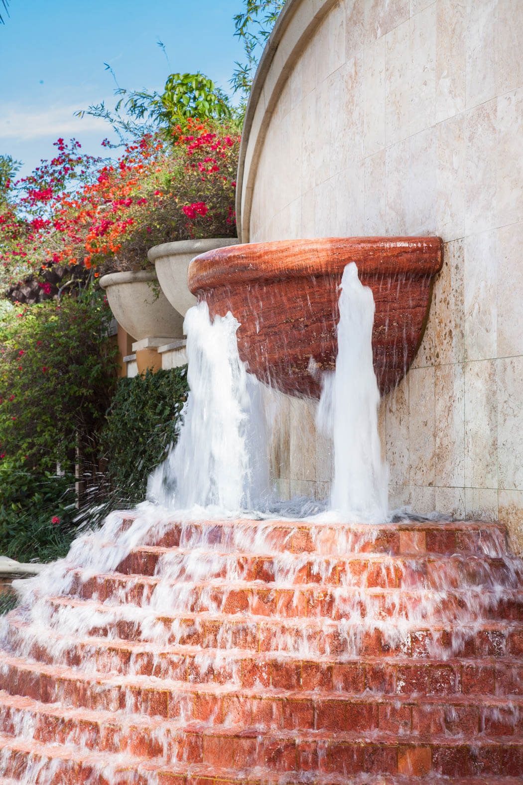 Brick fountain with water cascading down steps beside a white stucco wall and colorful flowers