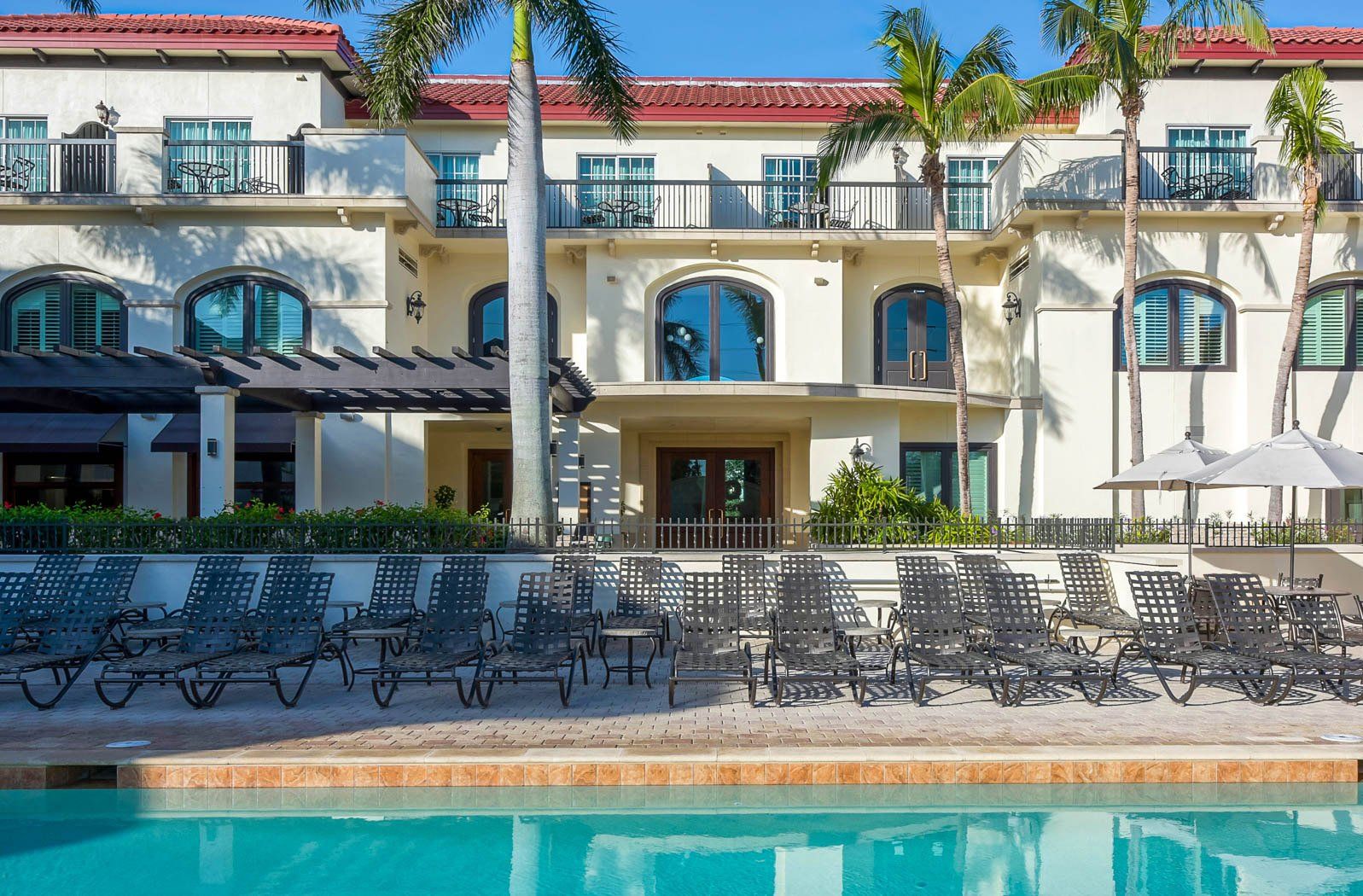Resort pool with lounge chairs, palm trees, and a Mediterranean-style building under a clear blue sky