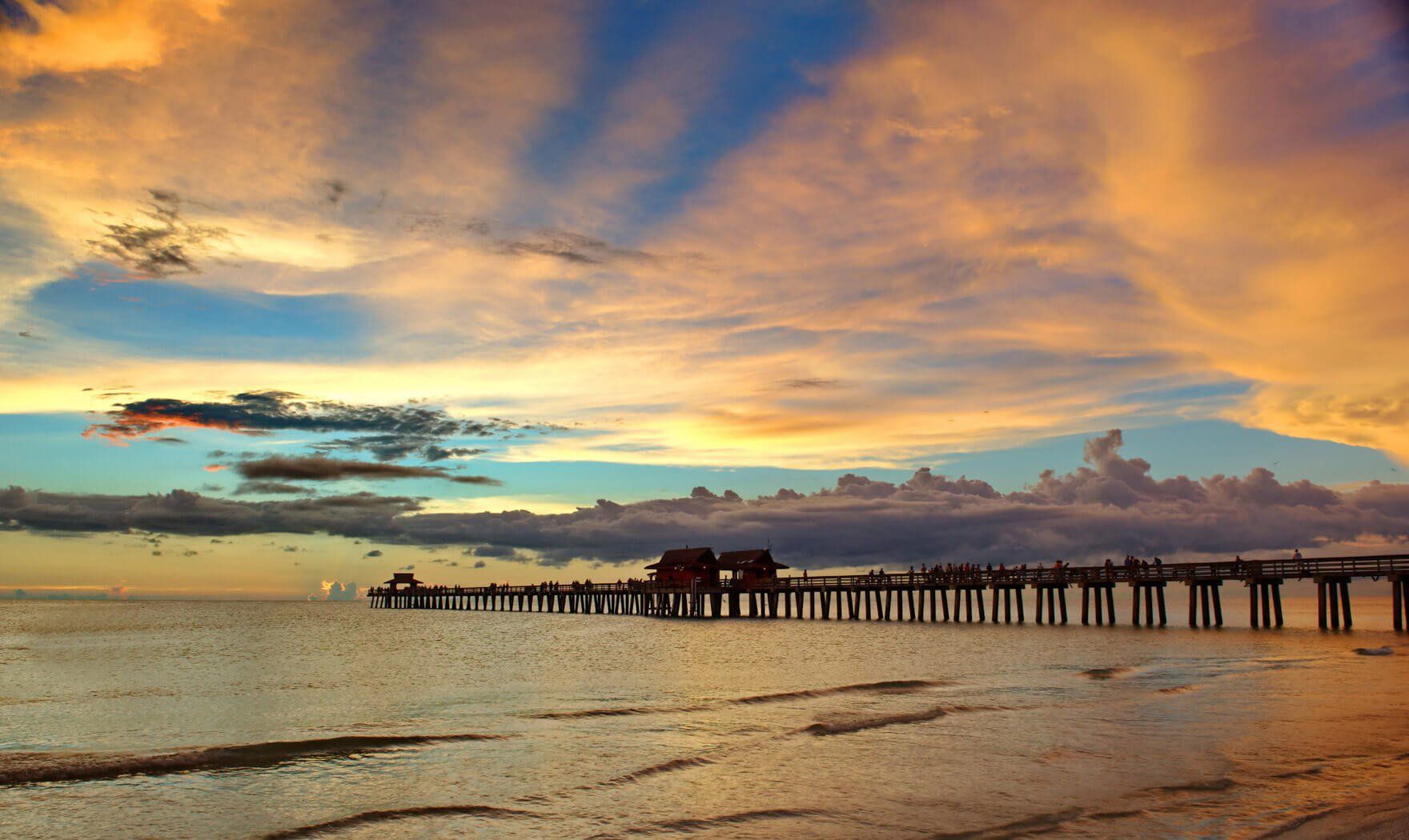 Sunset over a beach with a long wooden pier and colorful clouds reflected on wet sand