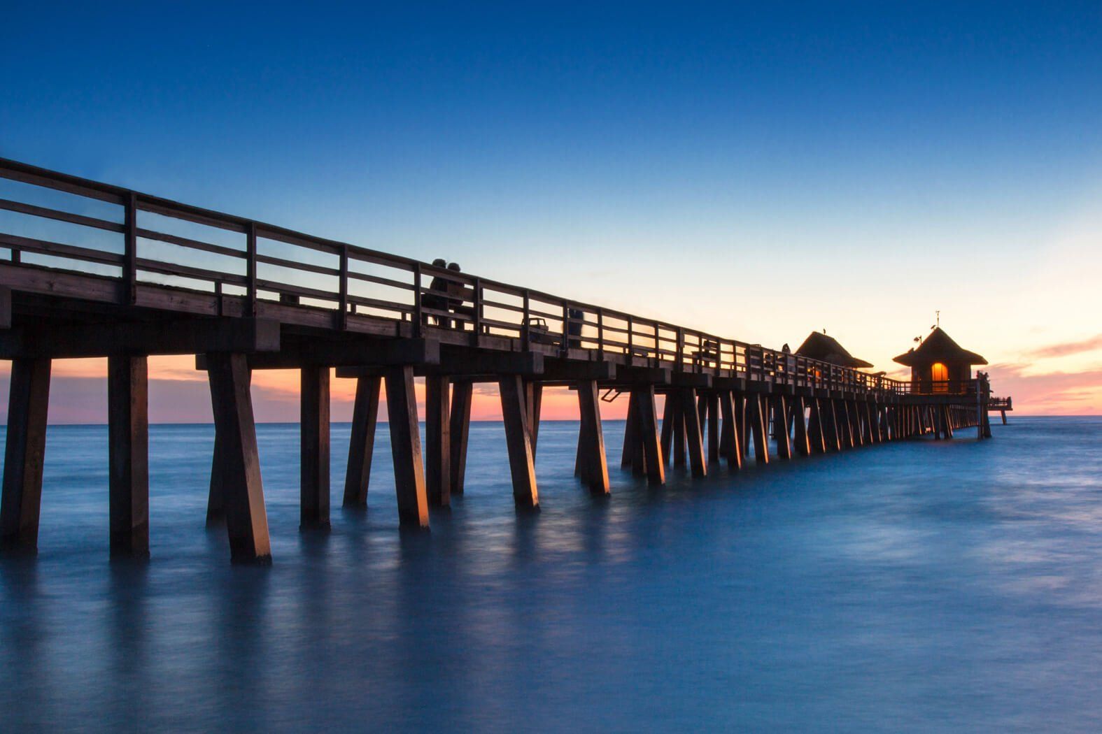 Wooden pier over calm blue water at sunset, leading to a small lit pavilion in the distance