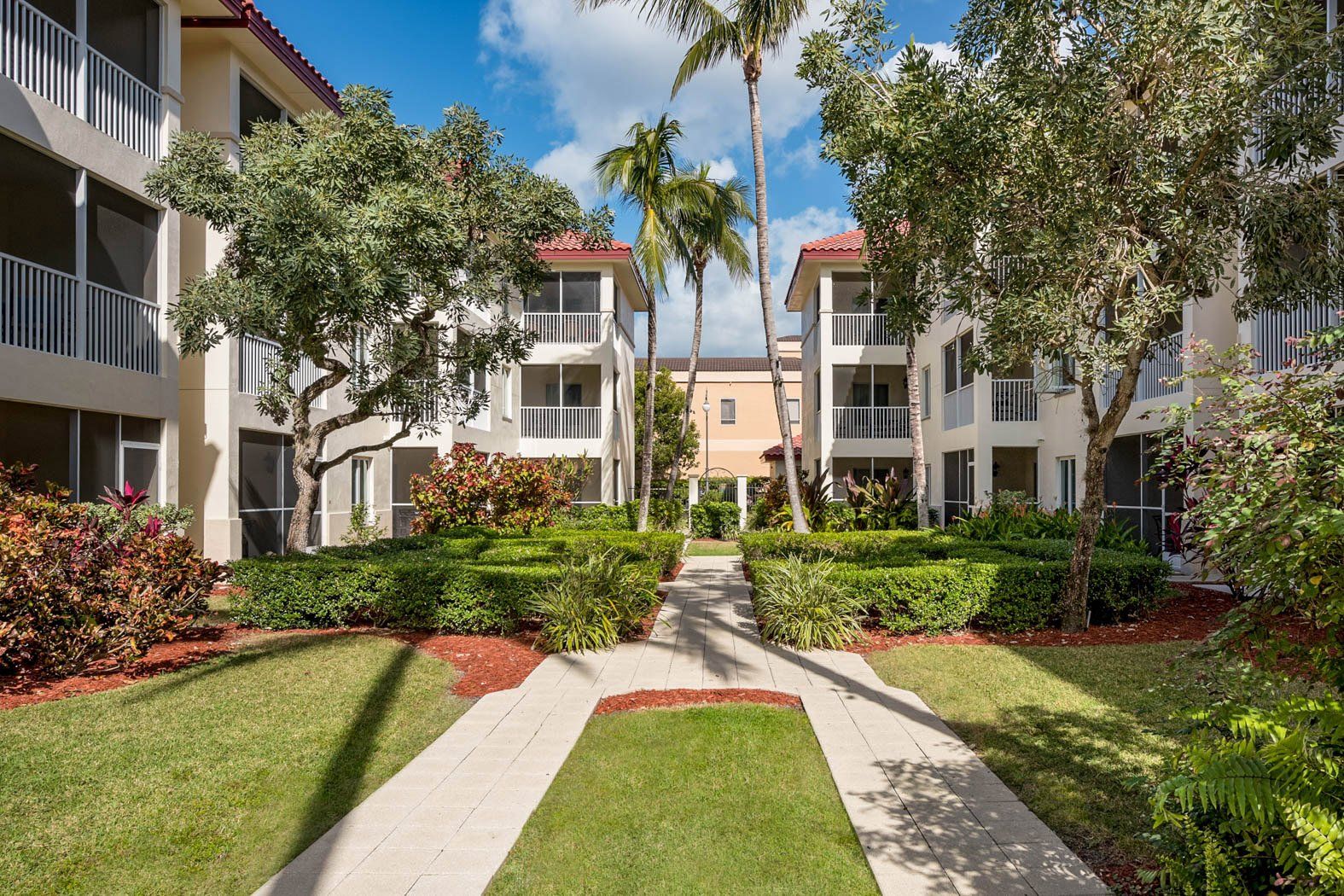 Courtyard path between white apartment buildings with palms, grass, and colorful shrubs under a blue sky