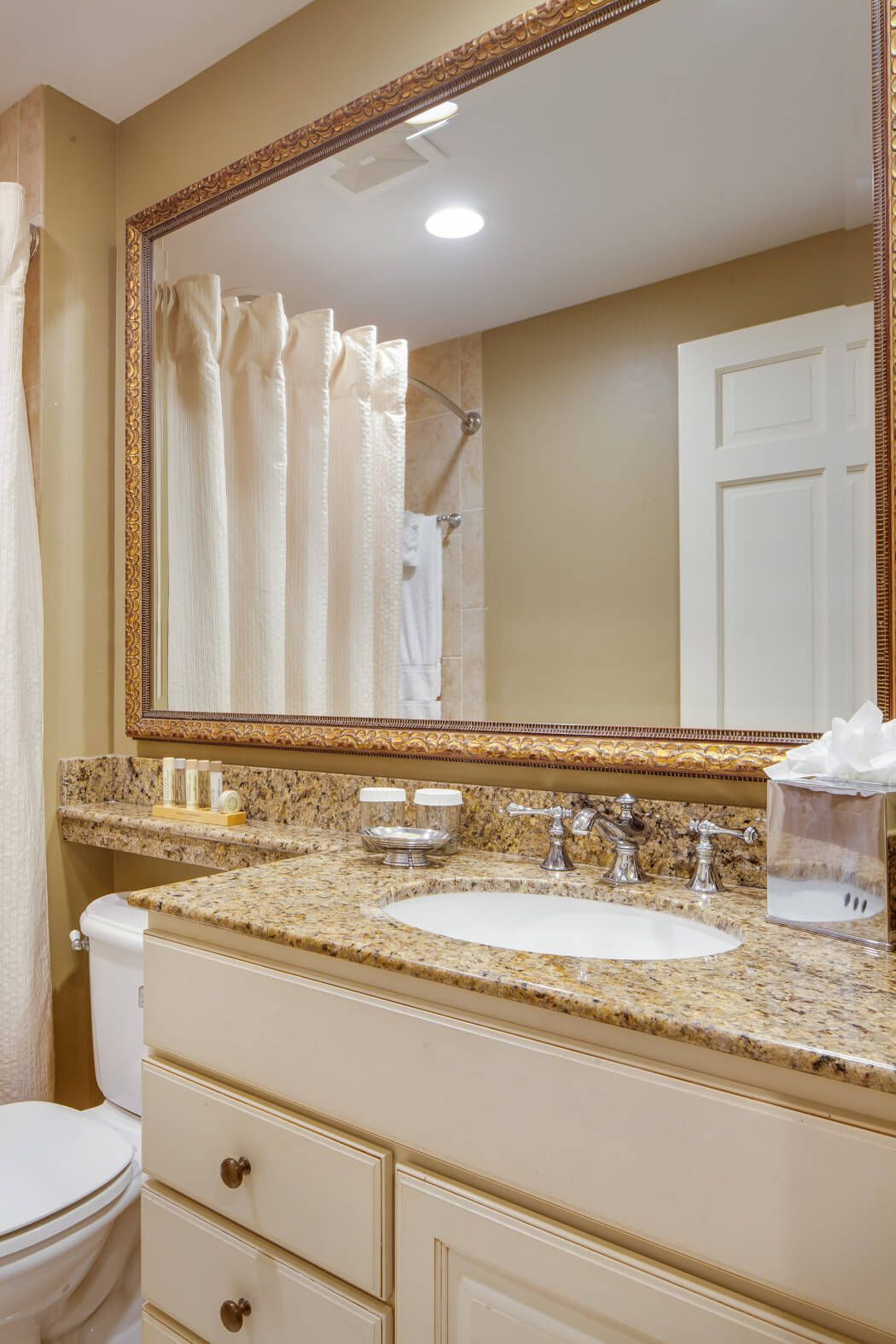 Bathroom vanity with granite countertop, white sink, gold-framed mirror, and beige tiled shower curtain