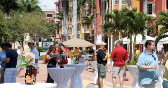 People mingling at an outdoor market on a sunny palm-lined street with tables and umbrellas.