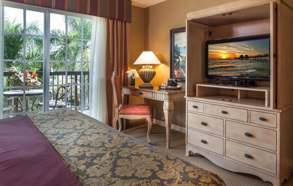 Hotel room with a bed, dresser TV, desk, lamp, and balcony view through sliding glass doors