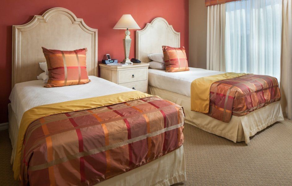 Hotel room with two twin beds, cream headboards, orange patterned bedspreads, and a bedside lamp by the window.