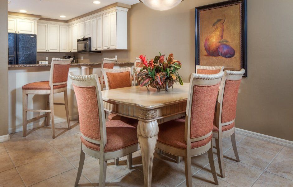 Dining room with a round table, pink chairs, flower centerpiece, and a kitchen in the background