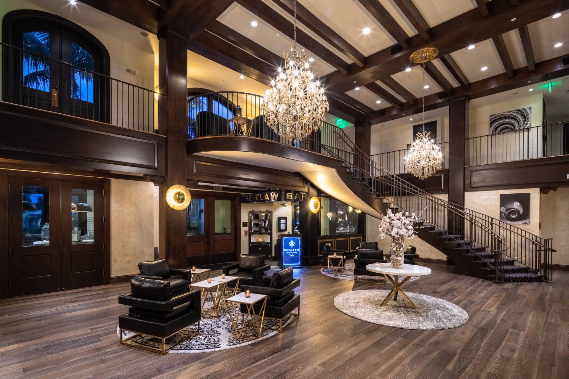 Grand foyer with chandeliers, dark wood beams, staircase, and seating area under vaulted ceiling.