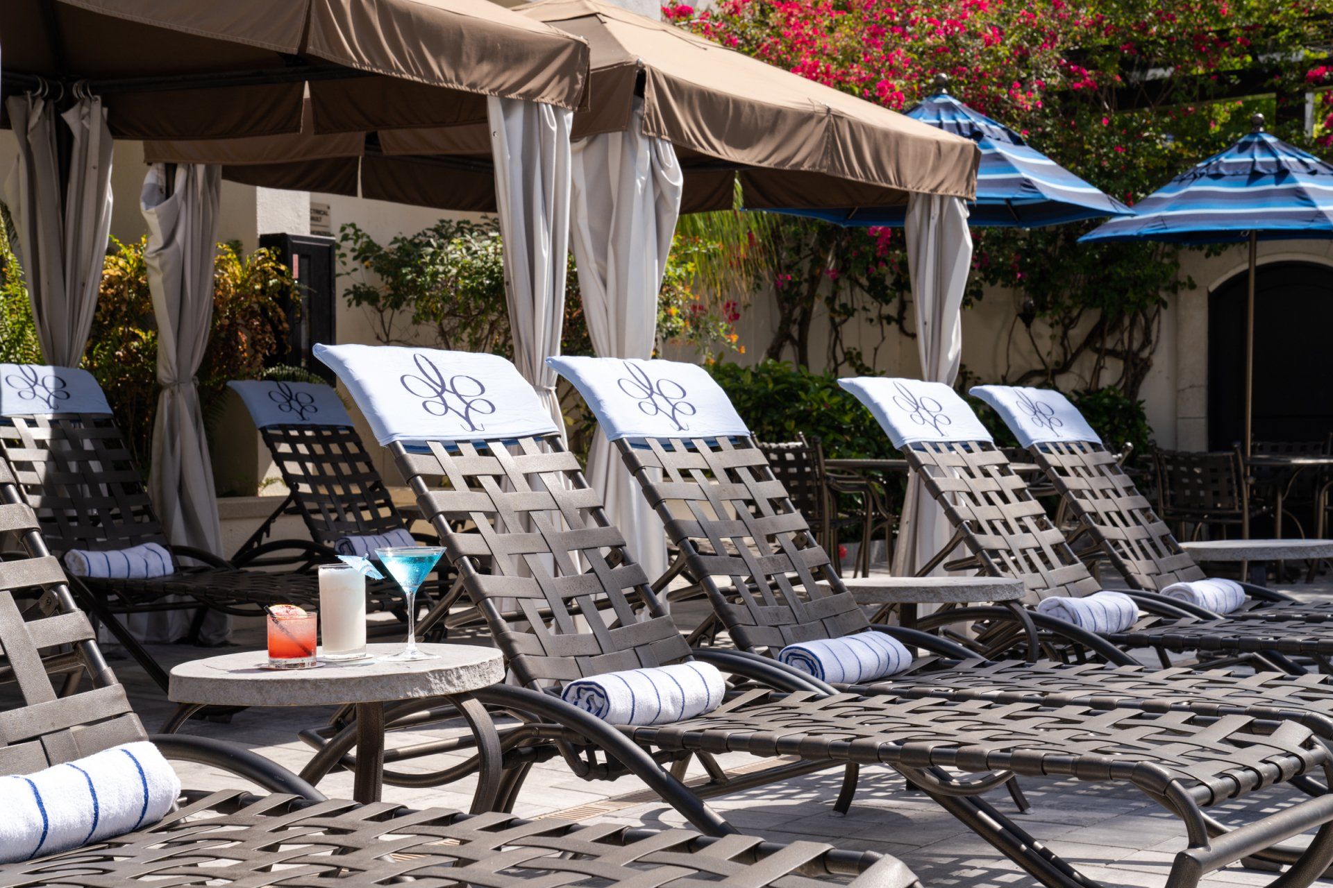 Row of empty black lounge chairs by a pool under beige and blue umbrellas
