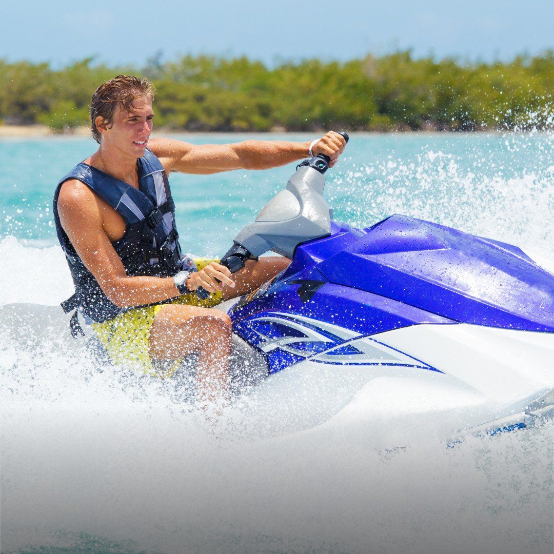 Person riding a blue-and-white jet ski, spraying water on a sunny day near a tropical shoreline