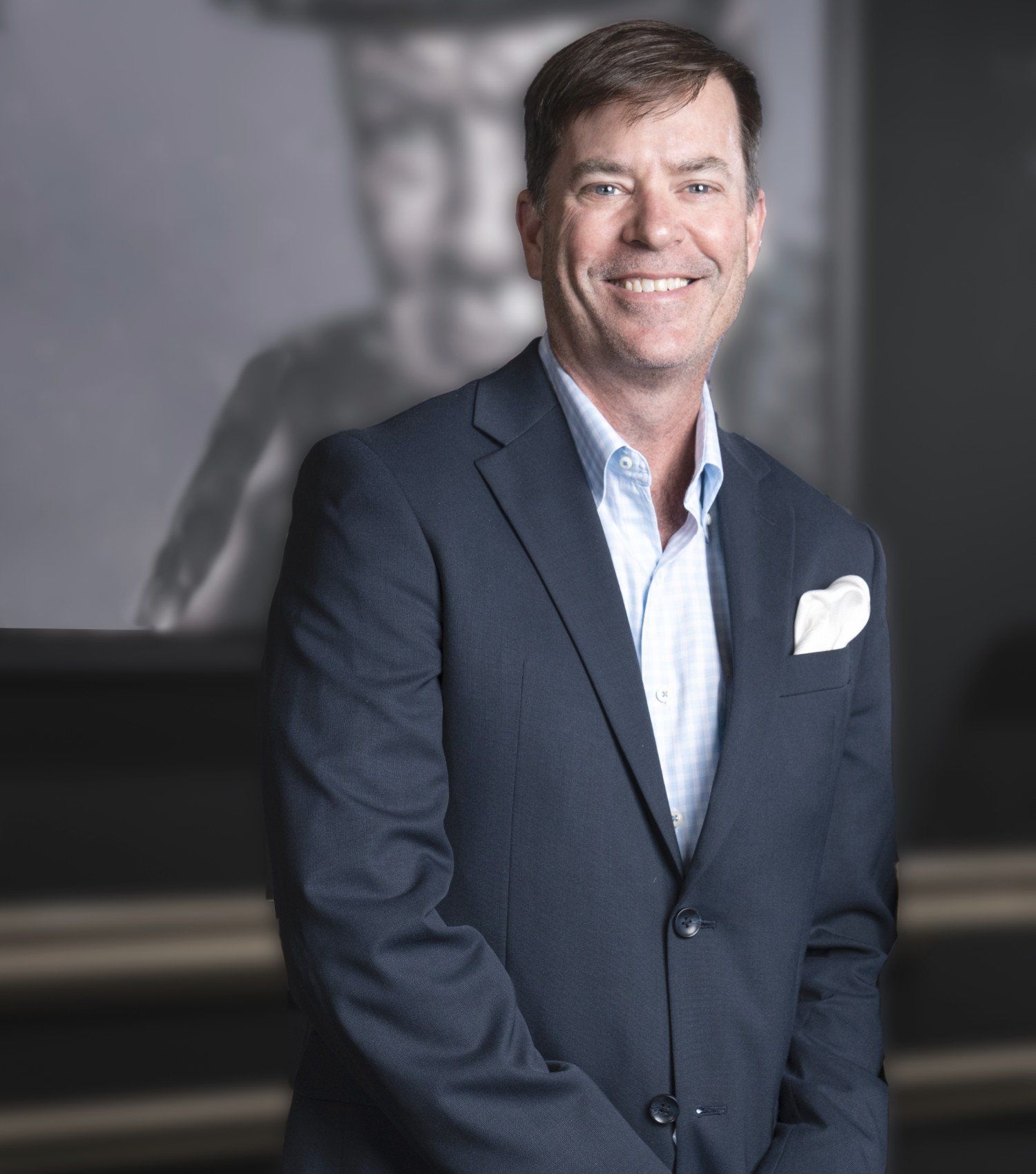 Smiling man in a dark blazer and light blue shirt standing indoors against a gray background.