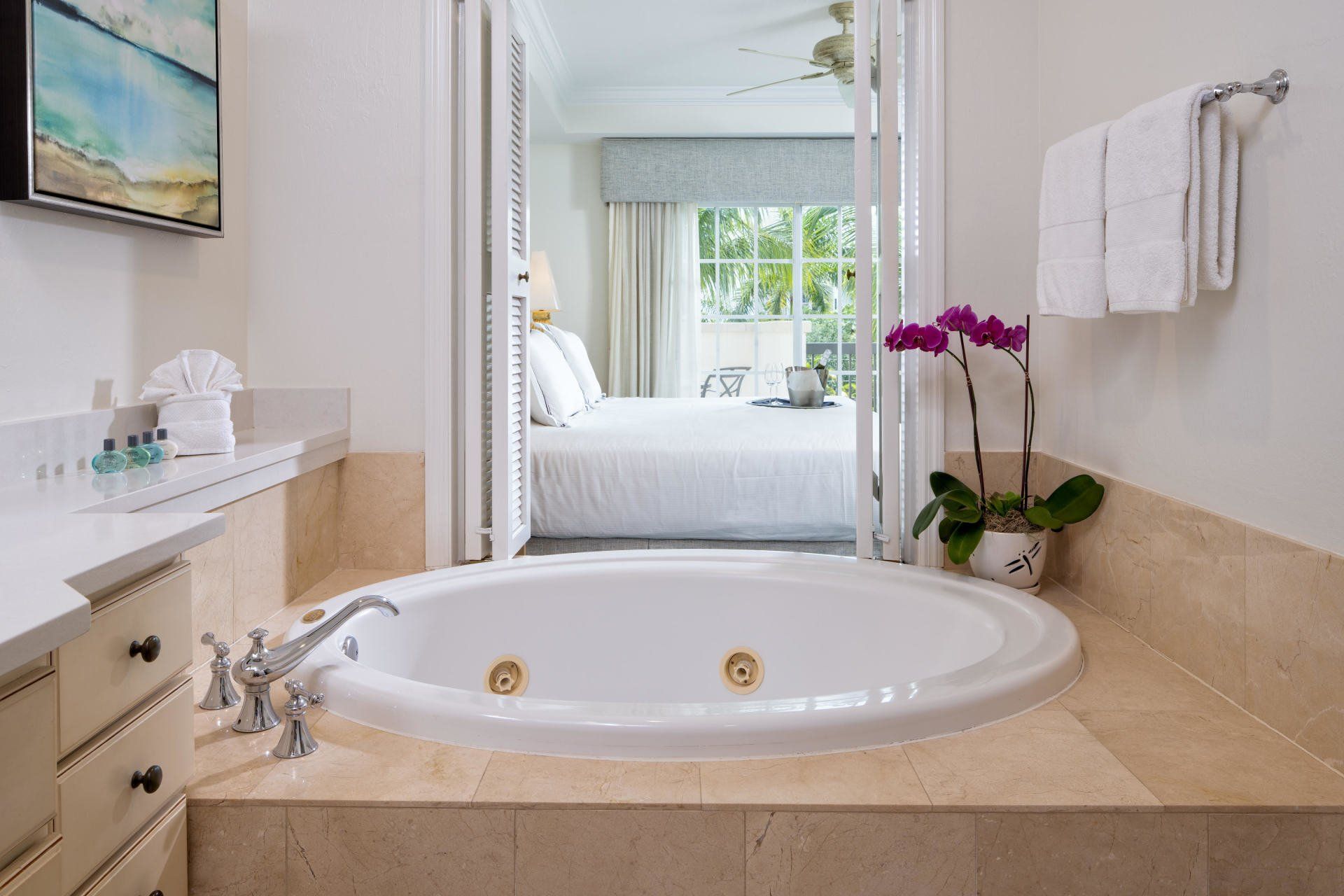 Bright bathroom with a corner soaking tub, beige marble, and a bedroom view beyond the glass doors