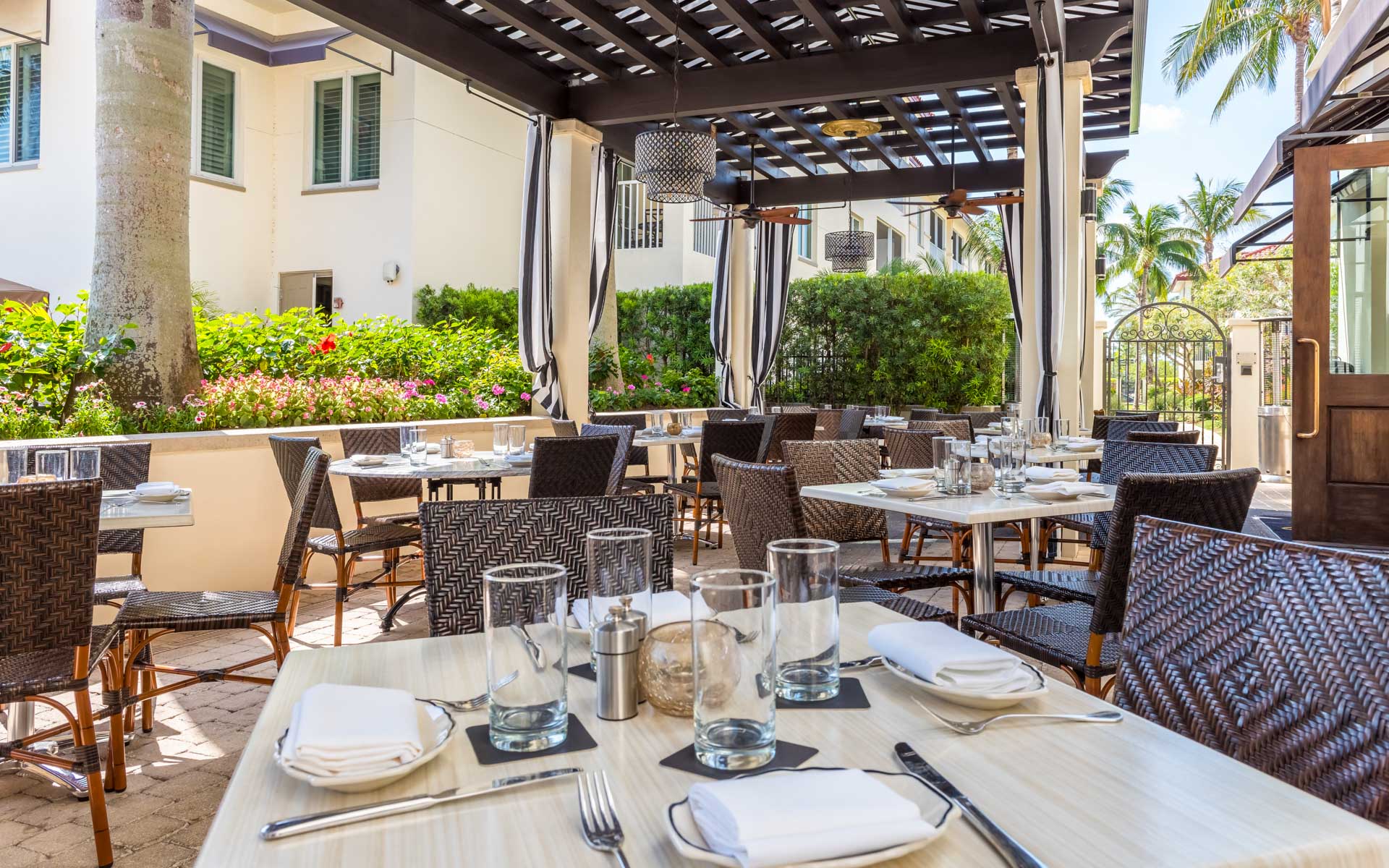 Outdoor restaurant patio with white tables, woven chairs, and a pergola in bright daylight