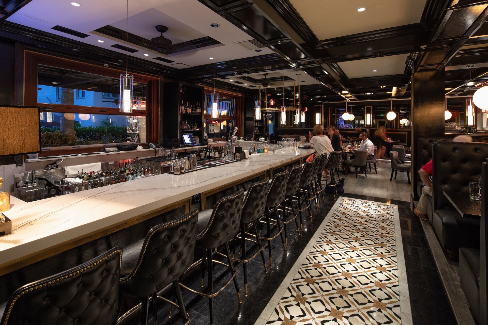 Empty modern restaurant bar with a long counter, black stools, and warm ambient lighting.