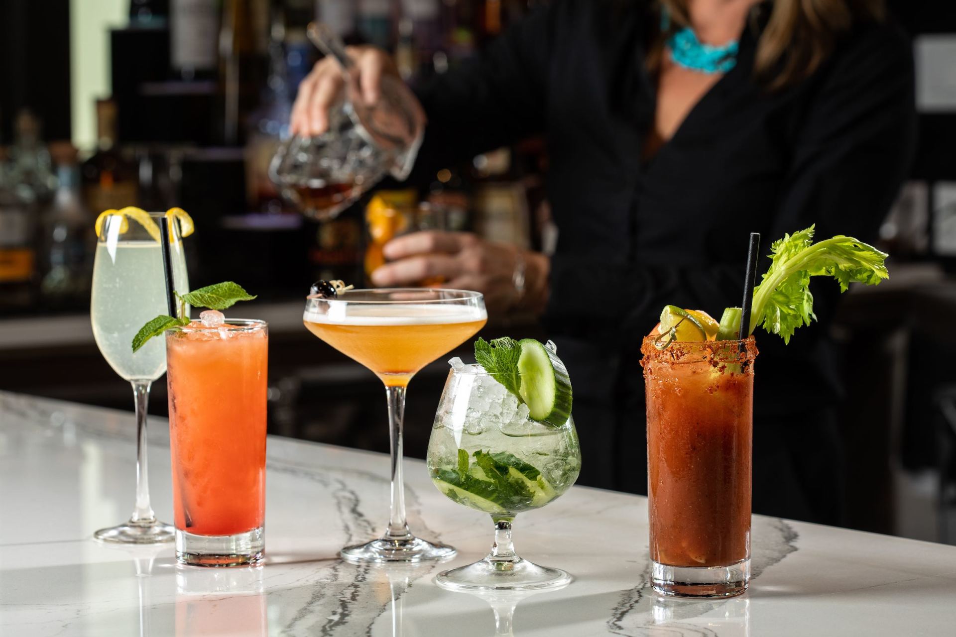 Five colorful cocktails on a bar counter, with a bartender pouring in the background.
