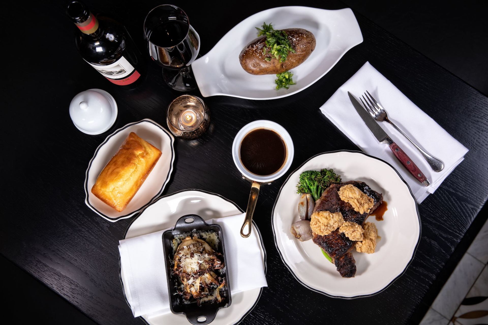 Top-down view of a gourmet meal with three plated dishes, wine, and utensils on a dark table