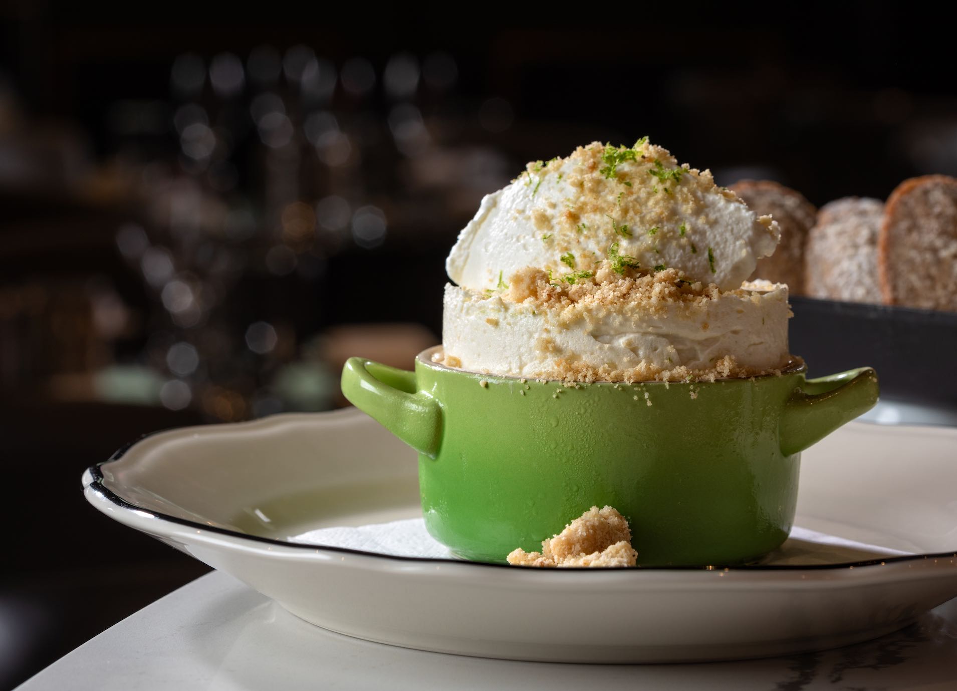 Green bowl with ice cream topped with crumbs, served on a white plate in a dim restaurant setting