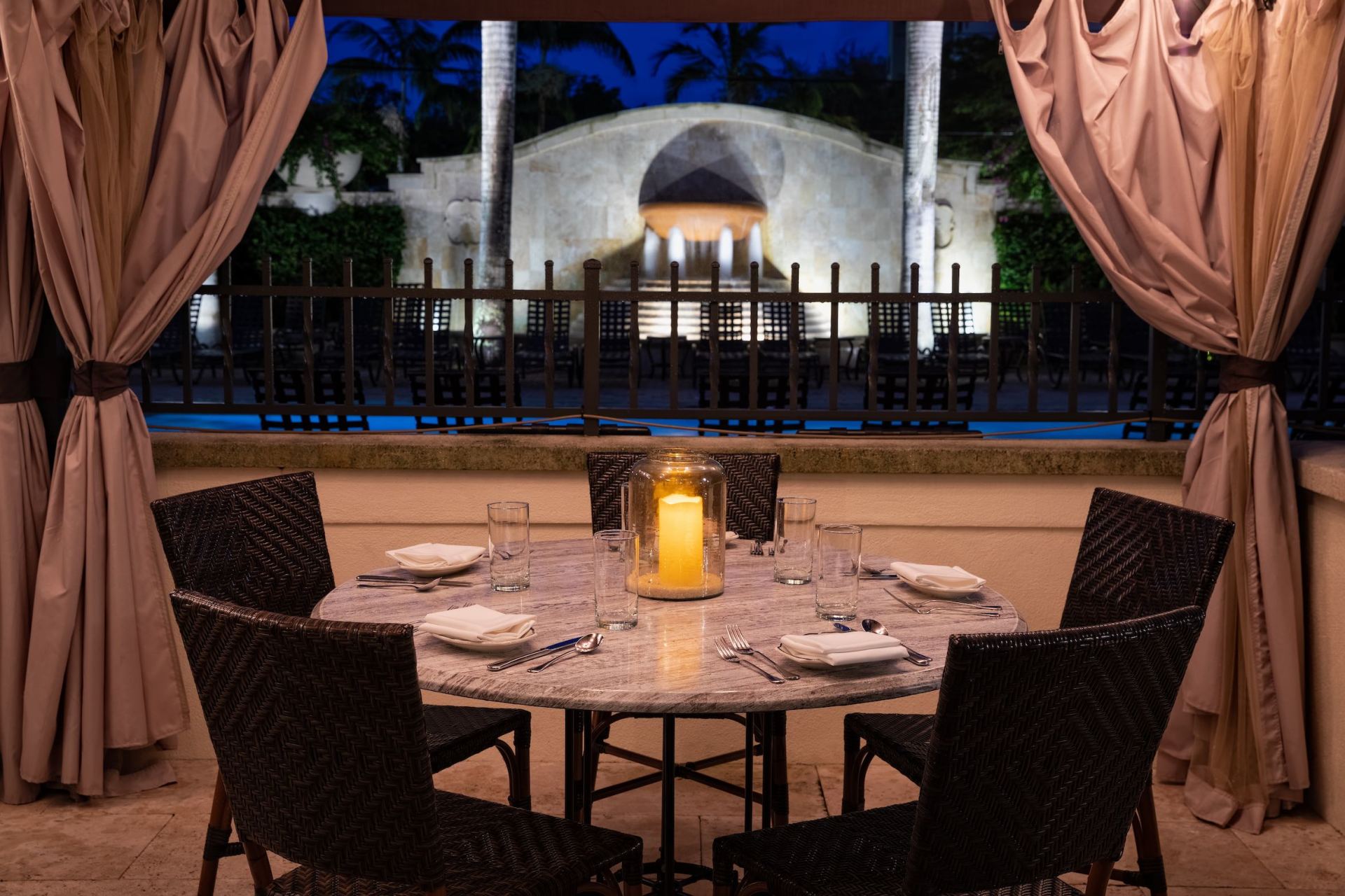 Candlelit patio table with dark chairs, set under draped curtains overlooking a lit pool at night