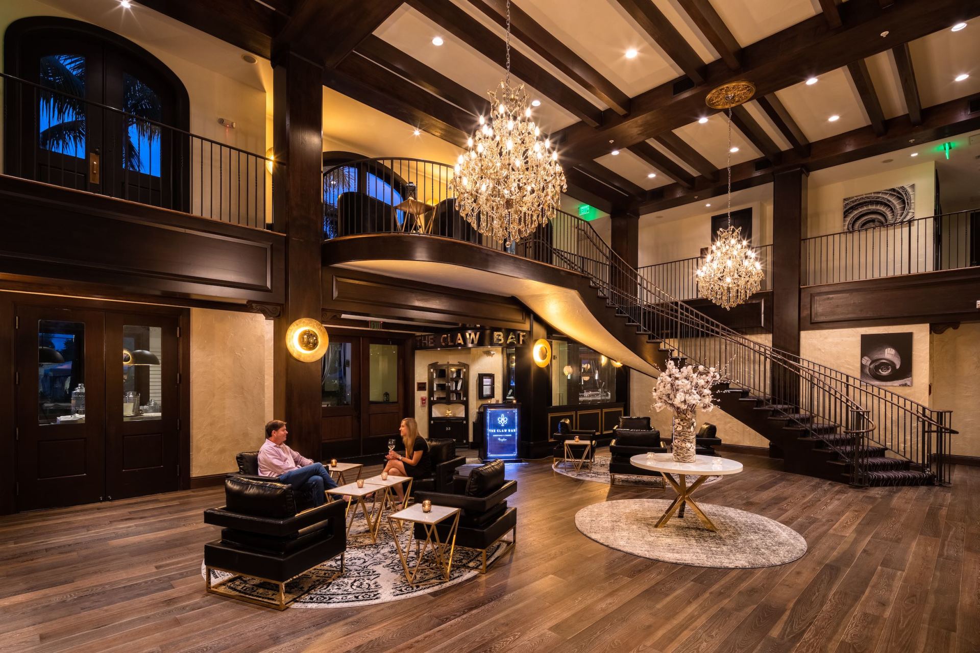 Grand hotel lobby with chandeliers, dark wood beams, a grand staircase, and a few seated guests.