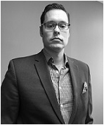 Homme en blazer et lunettes, regardant droit devant lui avec une expression neutre.