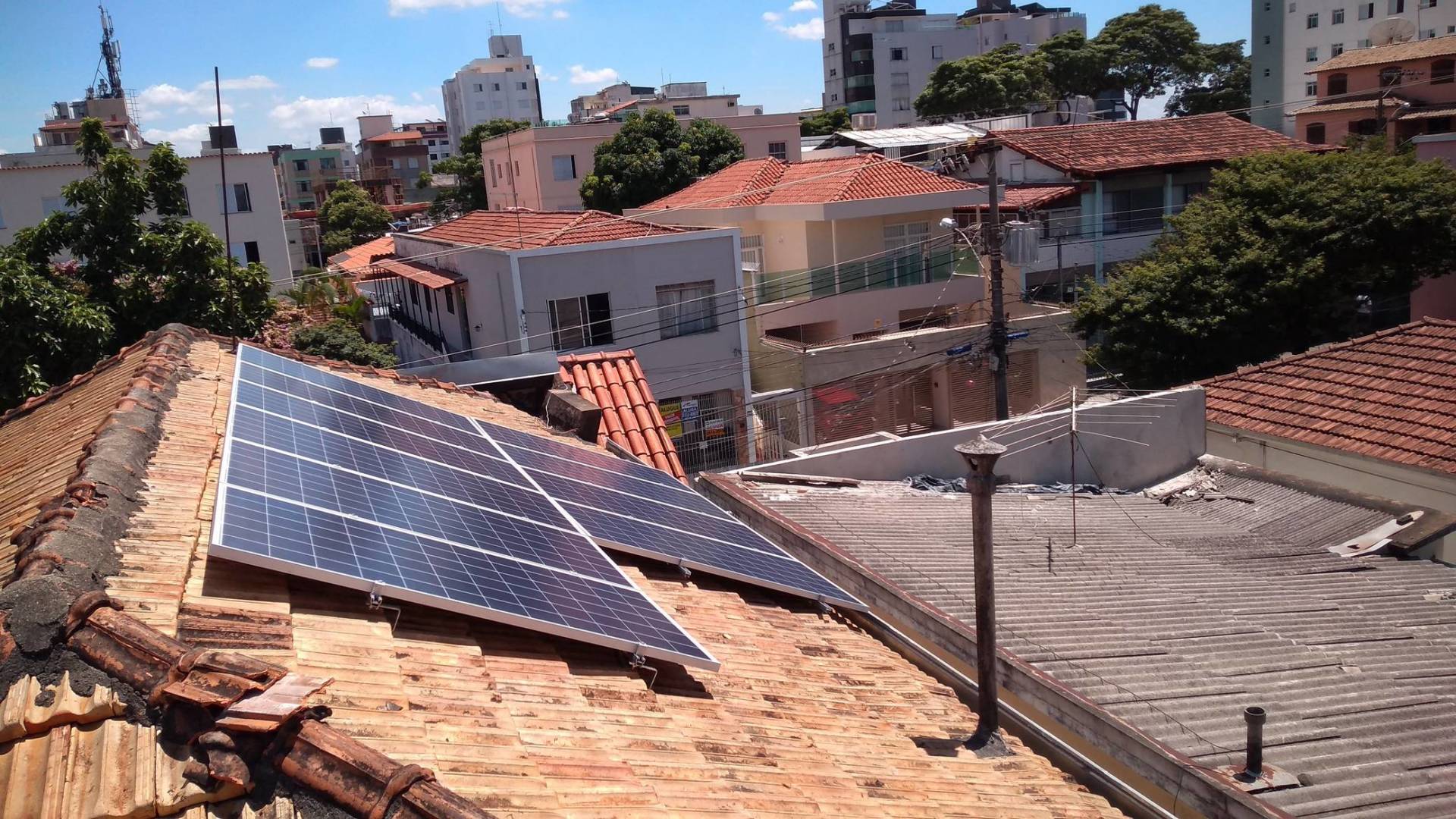 Painéis solares no telhado de telhas vermelhas com vista para um bairro da cidade sob um céu limpo.