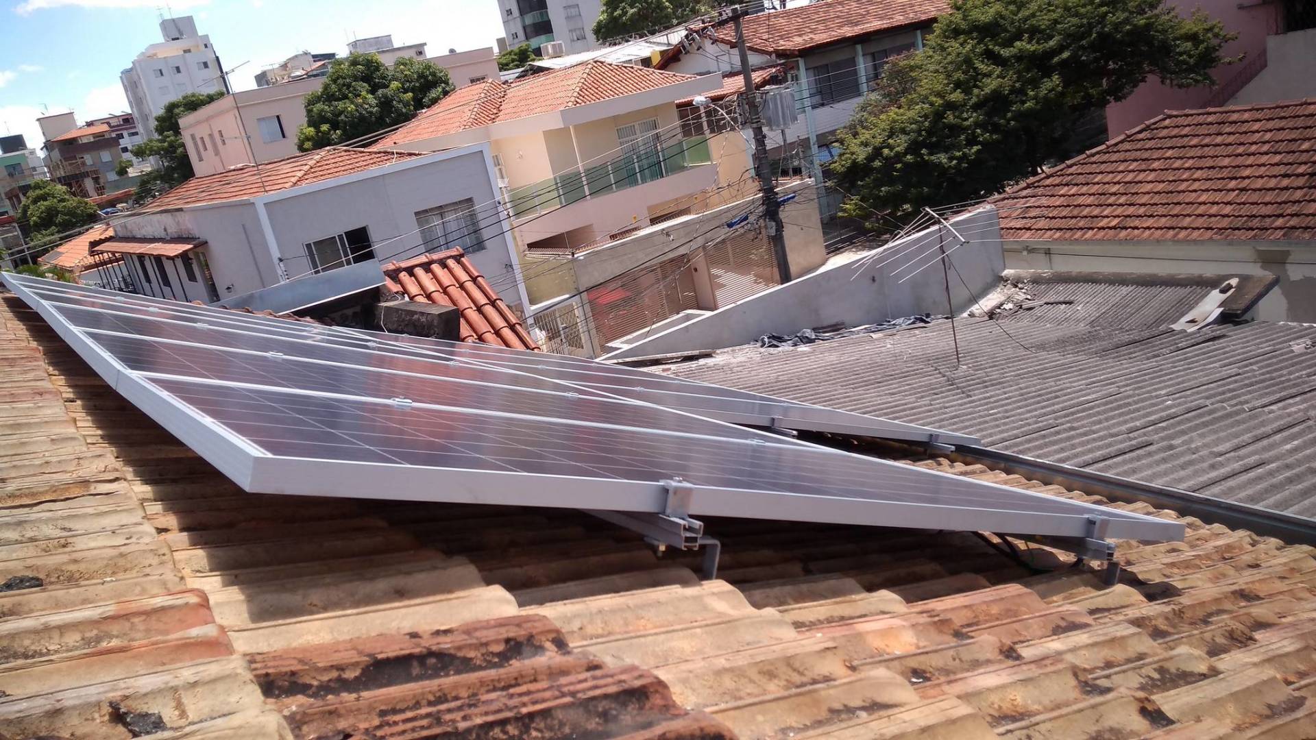 Painel solar inclinado instalado em um telhado, com casas e árvores vizinhas ao fundo.