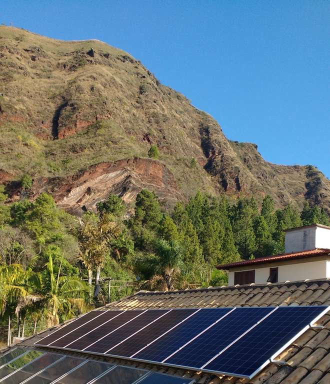 Painéis solares em um telhado com uma encosta rochosa e céu azul ao fundo.