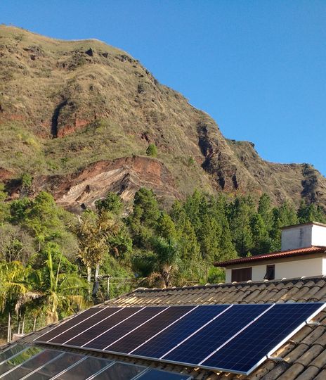 Painéis solares em um telhado sob uma encosta rochosa verdejante e um céu azul límpido.