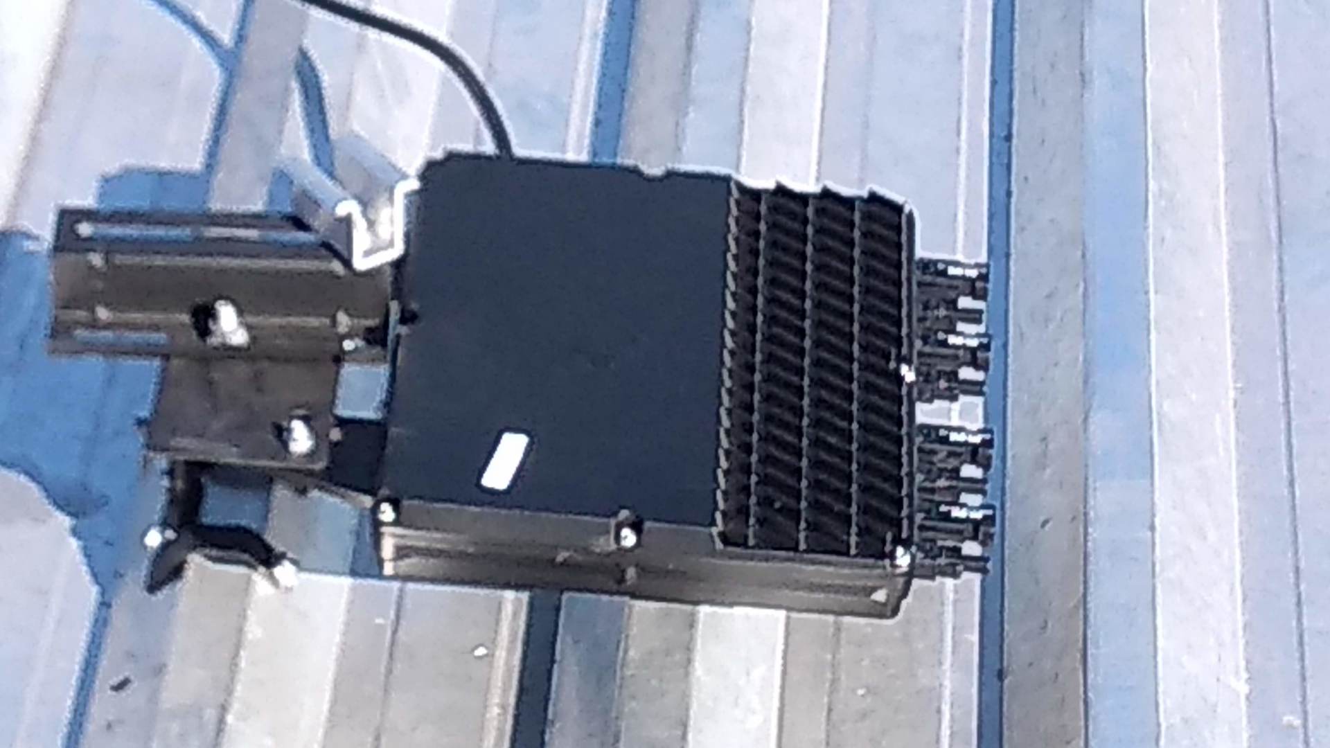 Micro inversor sobre uma superfície metálica ondulada, com uma extremidade aletada e cabos conectados.