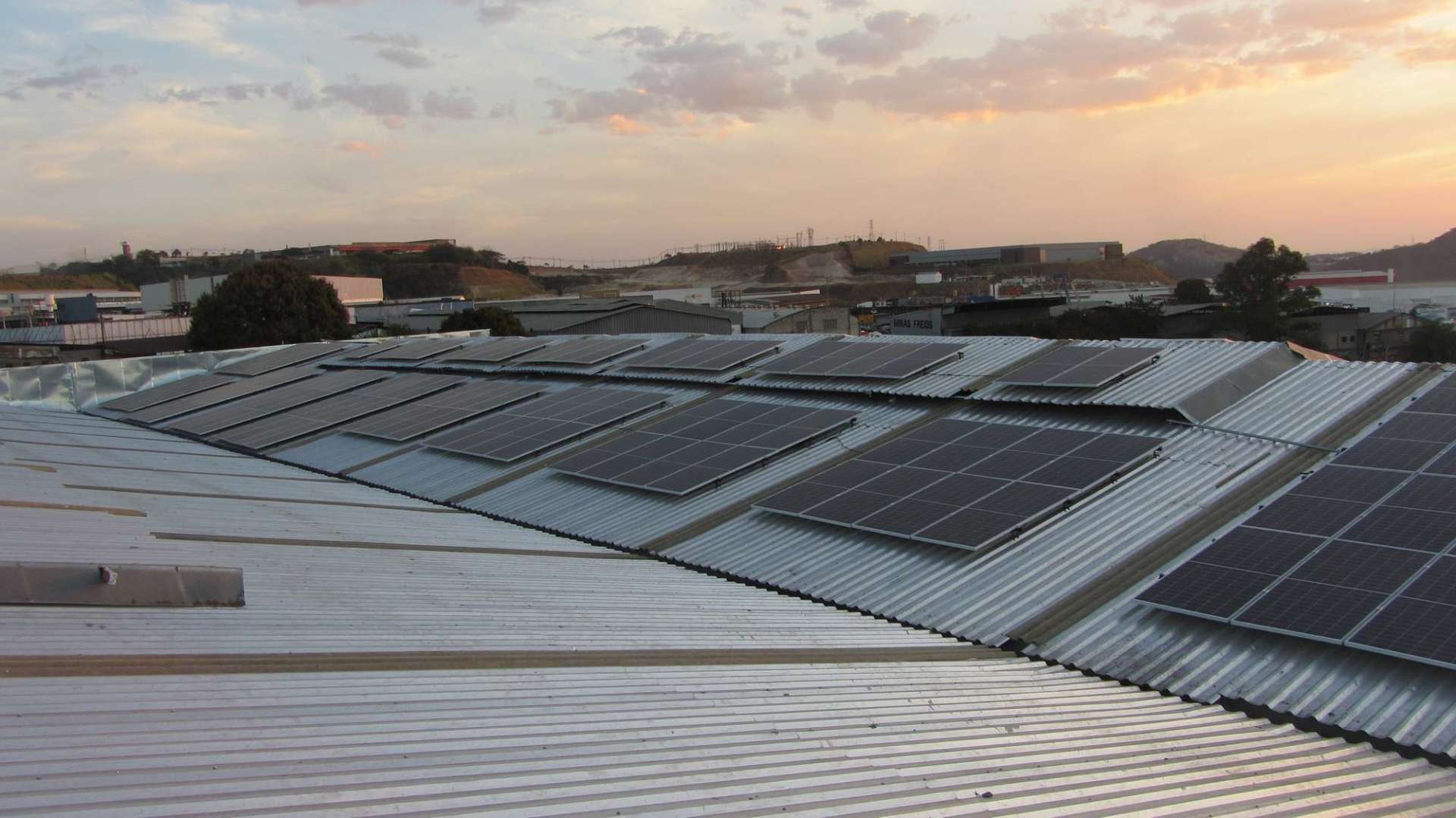 Painéis solares cobrindo um telhado ao pôr do sol, com prédios próximos e o horizonte da cidade ao fundo.