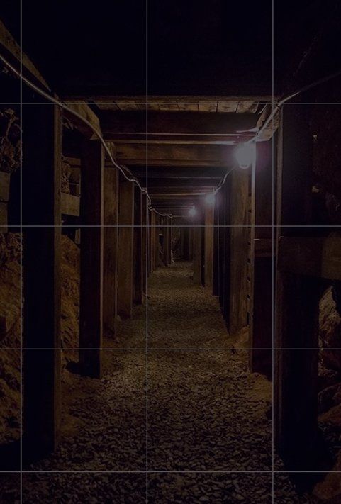 A dark, narrow underground tunnel with wooden support beams along the walls and a gravel floor, lit by small lamps.