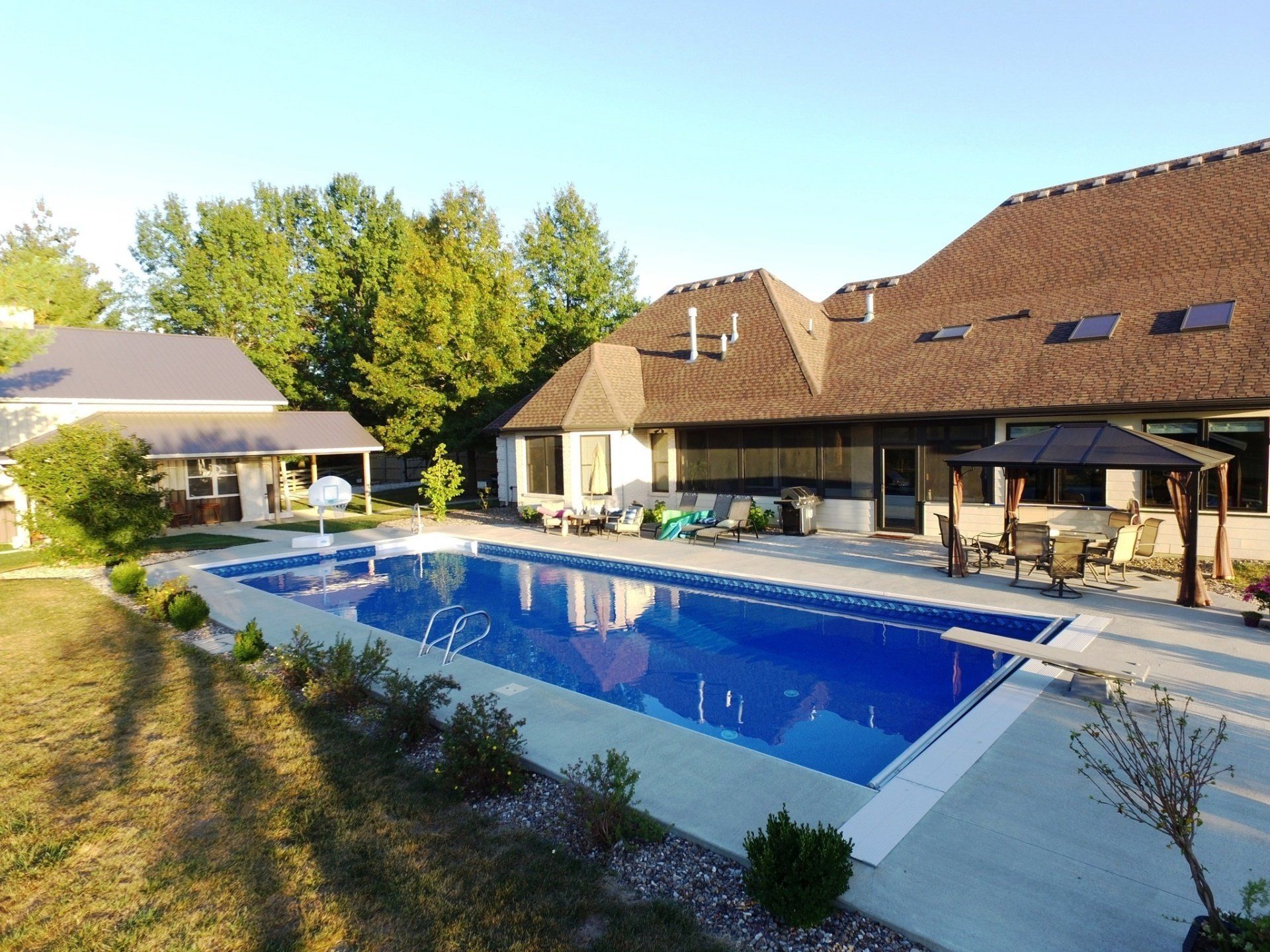 Backyard with a large blue swimming pool beside a house and patio, surrounded by trees and shrubbery