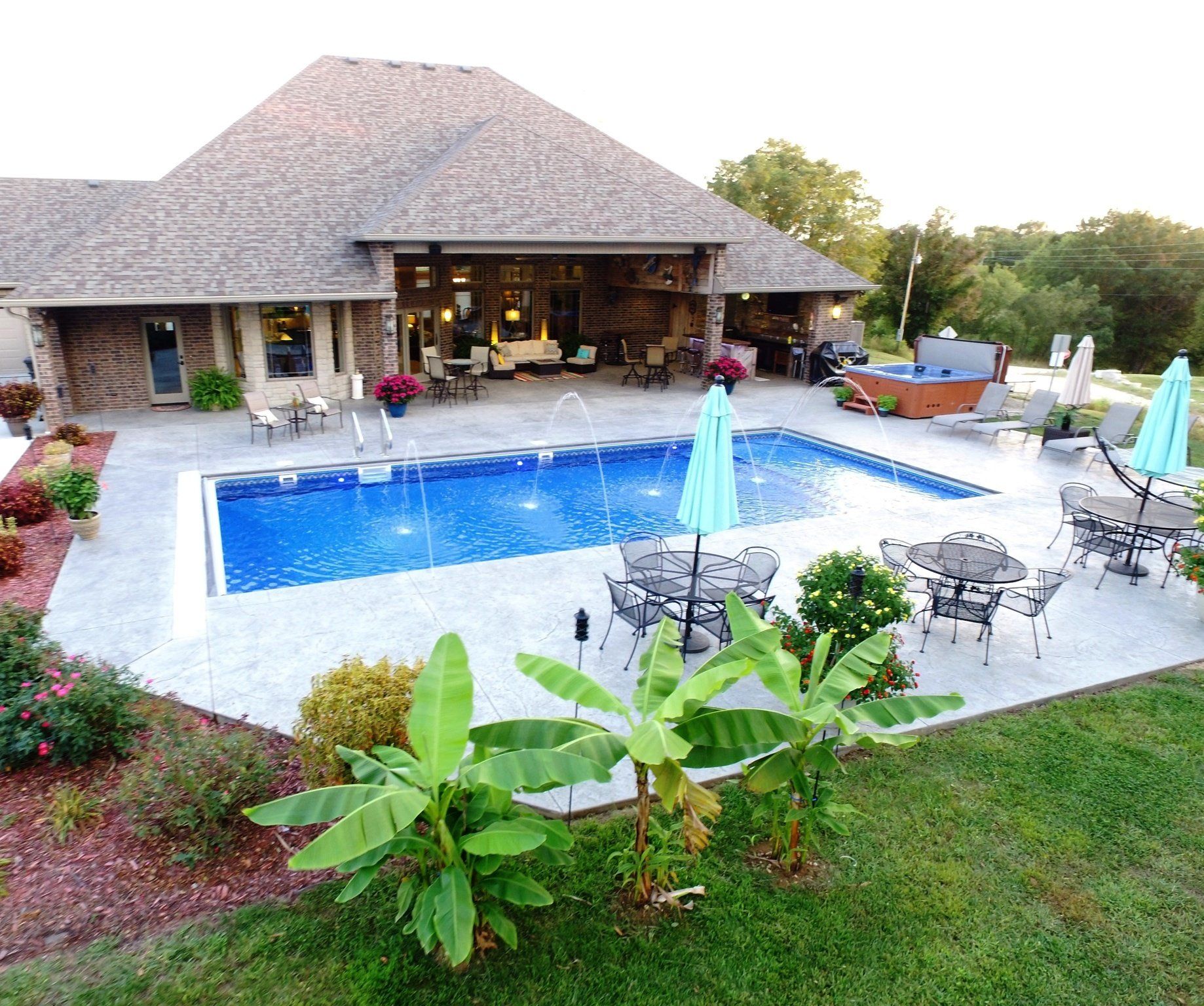 Backyard patio with a blue swimming pool, umbrellas, lounge chairs, and a stone house at dusk