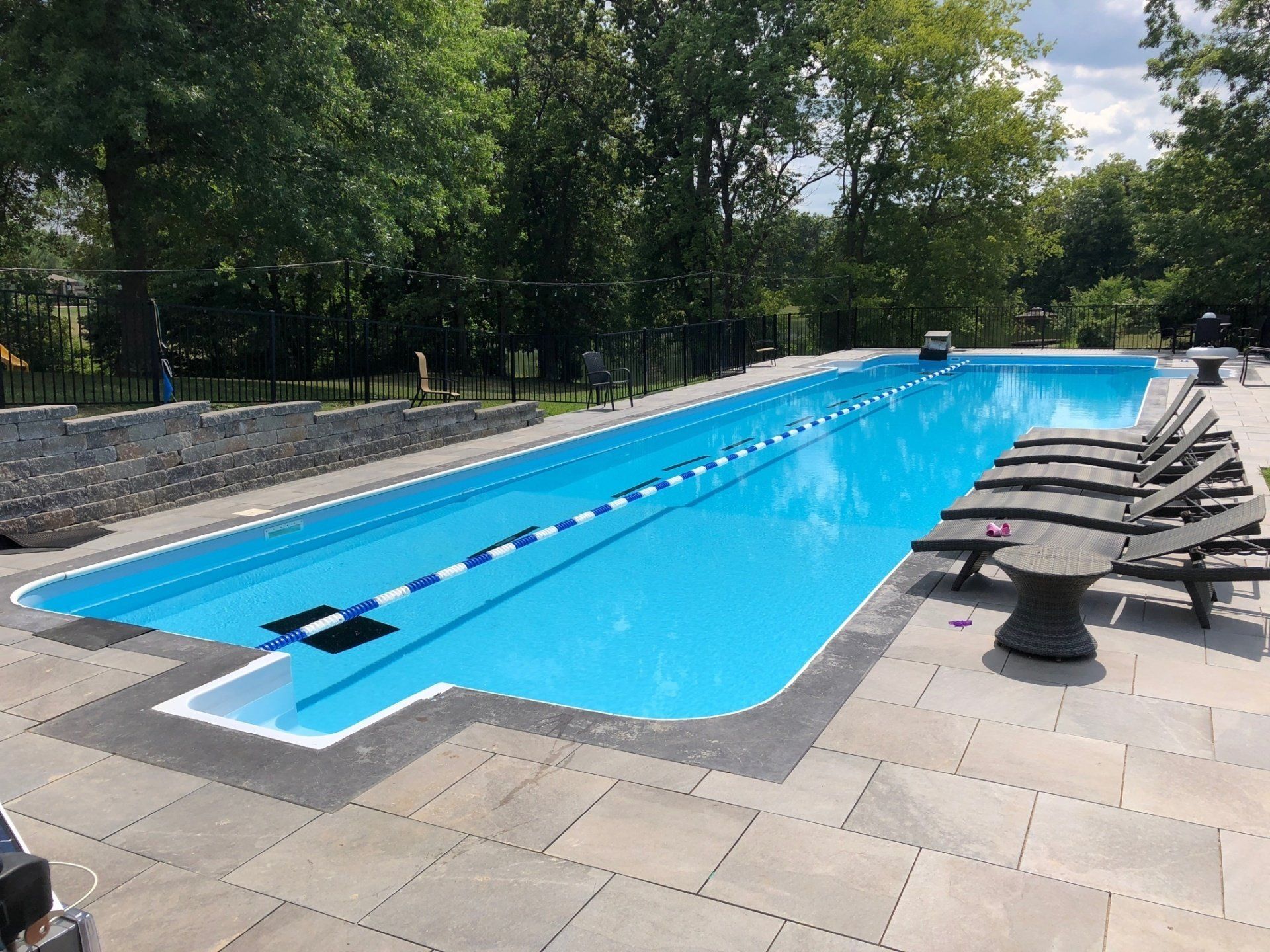 Outdoor lap pool with lane dividers, blue water, and lounge chairs on a stone deck surrounded by trees