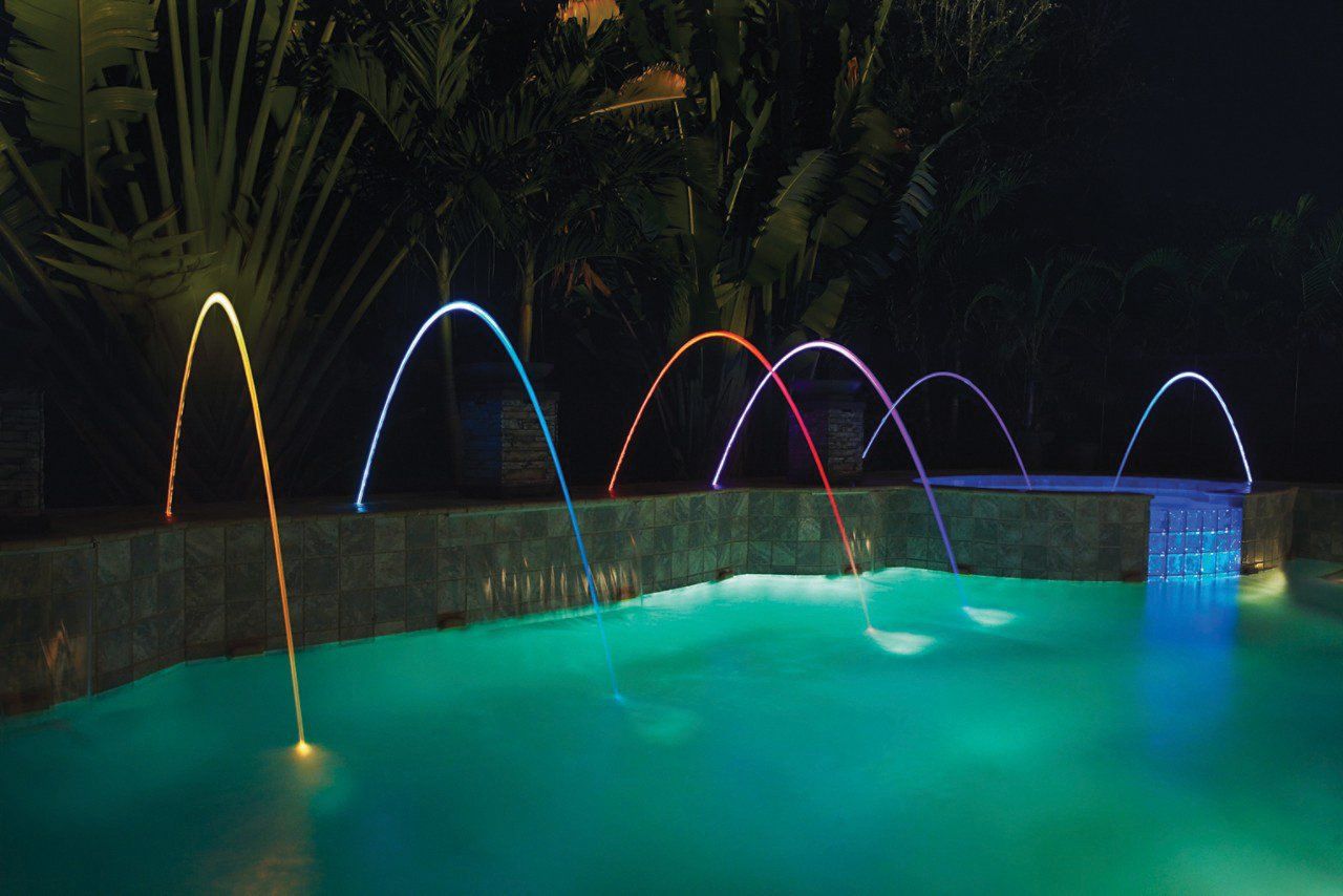 A swimming pool is lit up at night and has a waterfall in the background.