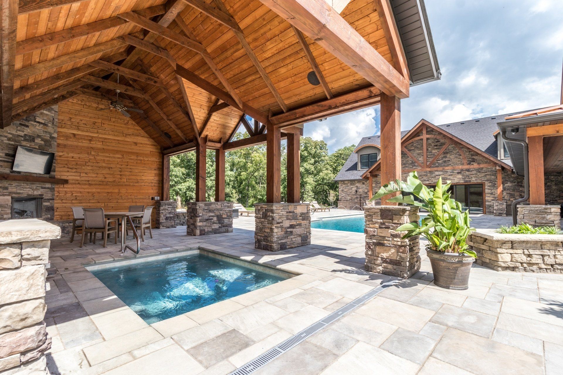 Covered stone patio with hot tub and pool beside a wooden pavilion
