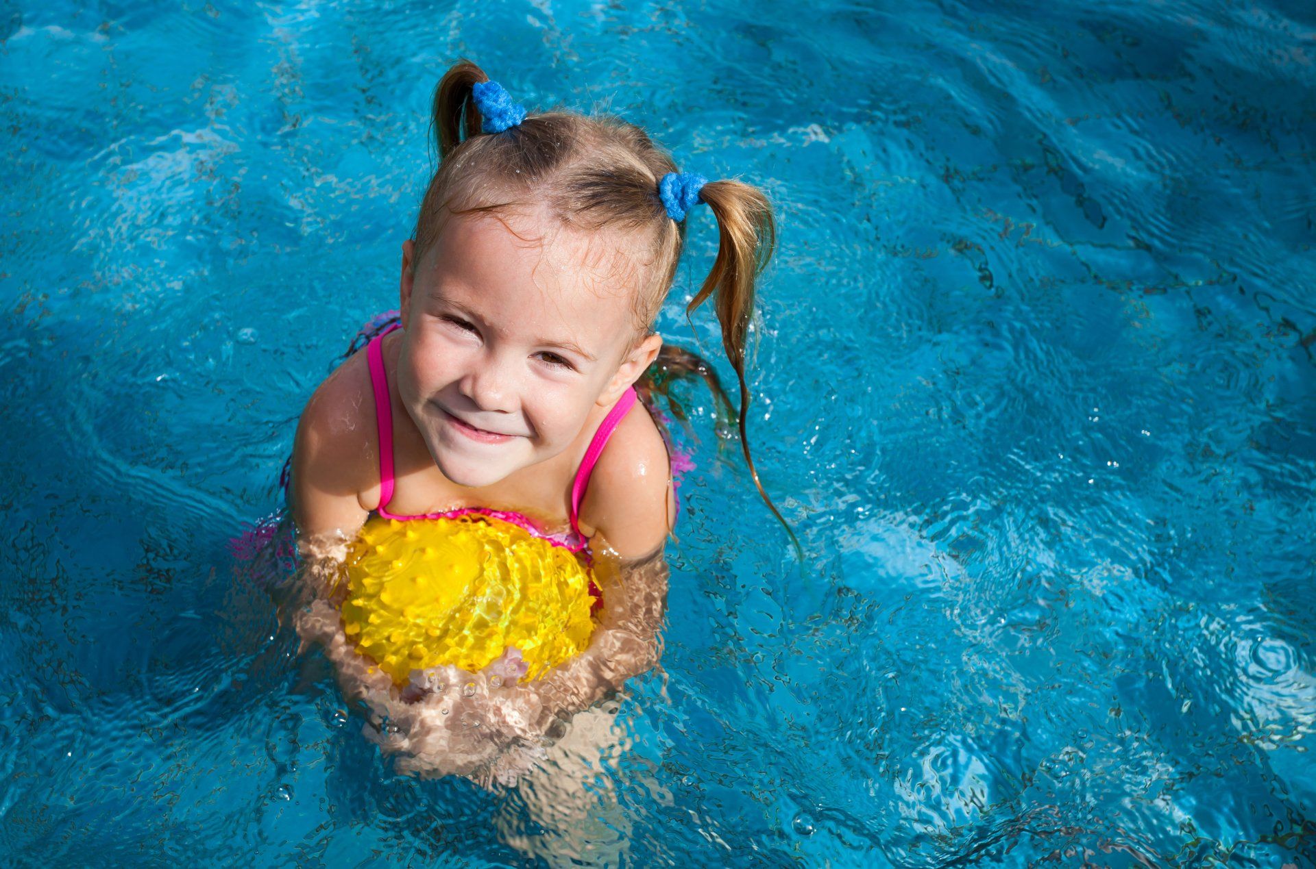 Smiling child in a blue swimming pool, holding a yellow toy and wearing pink goggles