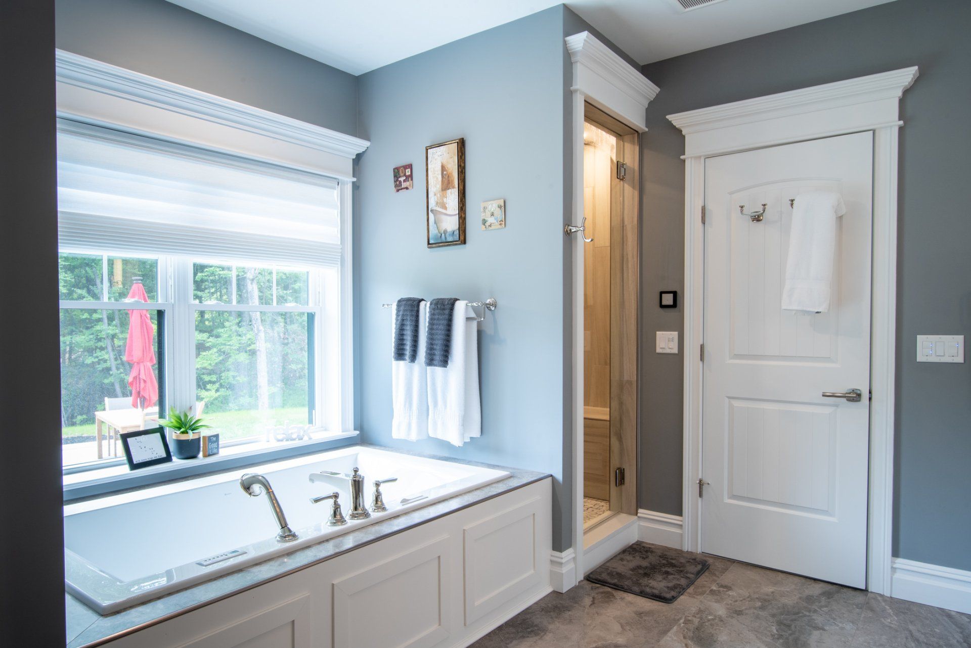 Bright bathroom with a white soaking tub, window, vanity, and shower door on gray walls