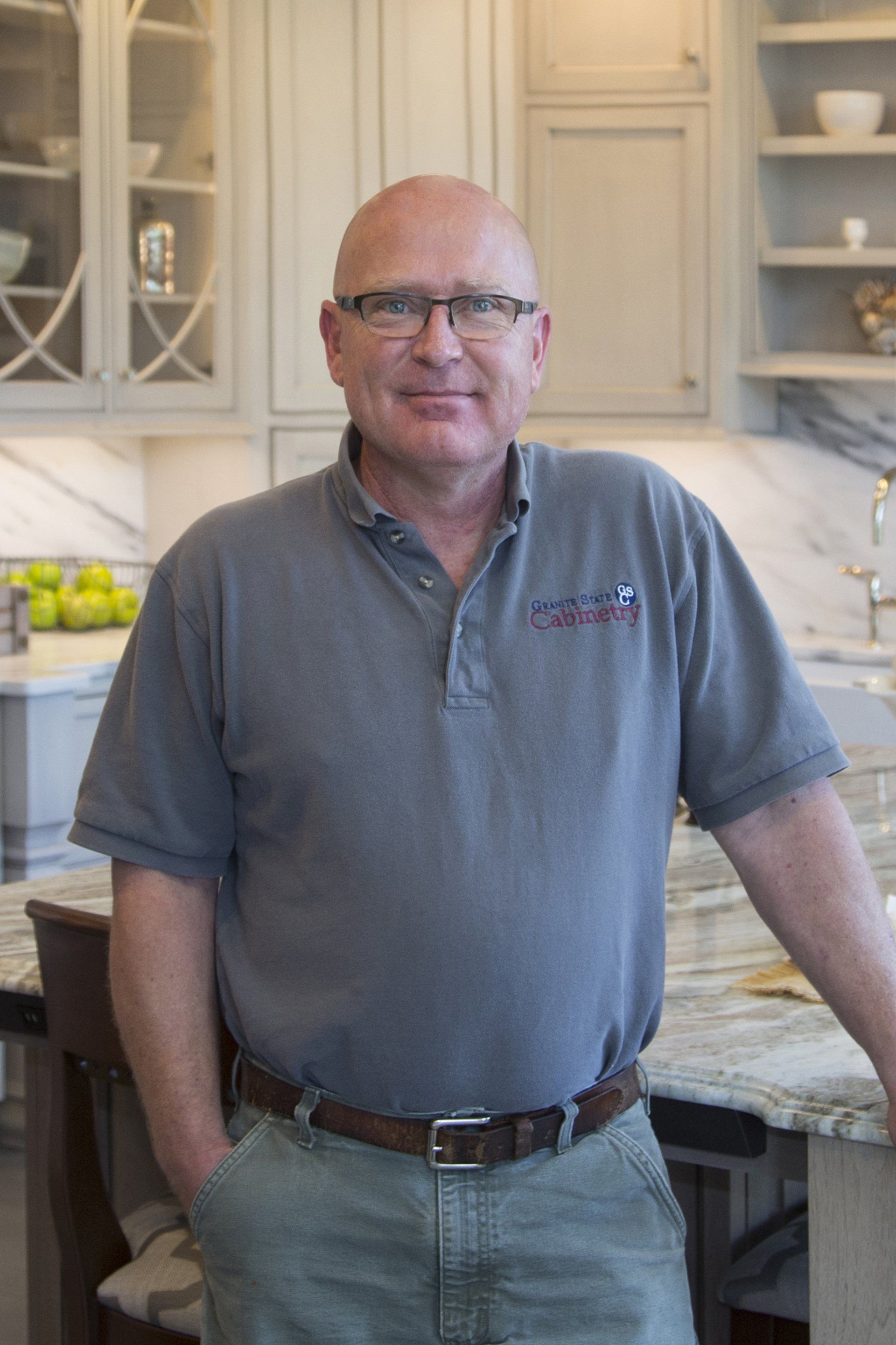 Man standing in a bright kitchen, wearing glasses and a gray polo shirt, smiling slightly.