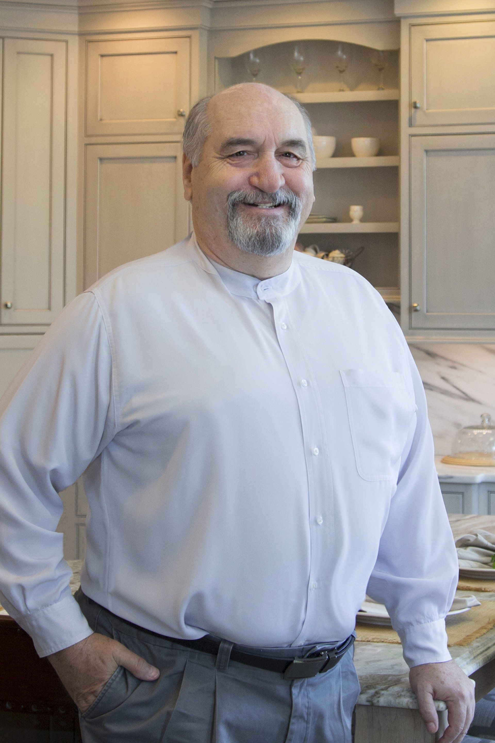 Man in a light button-up shirt standing in a kitchen with cream cabinets, smiling at the camera