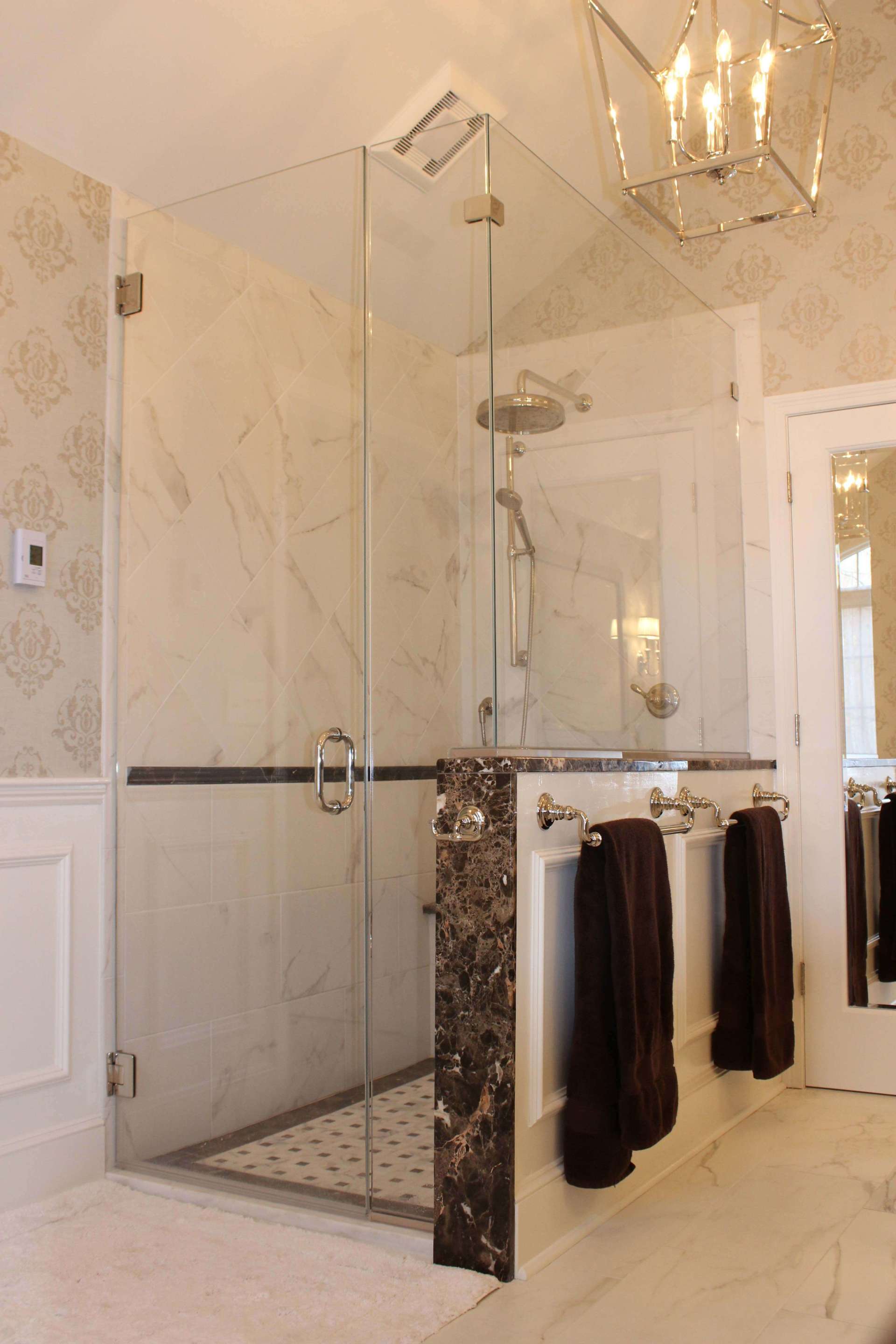 Elegant marble bathroom with glass shower enclosure, ornate chandelier, and hanging dark towels near a vanity.
