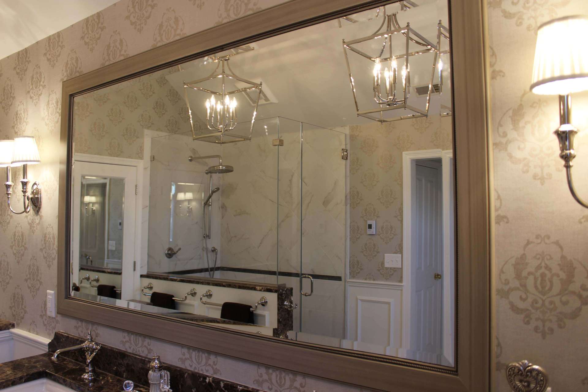 Elegant bathroom with large framed mirror, chandelier lights, marble vanity, and beige patterned wallpaper