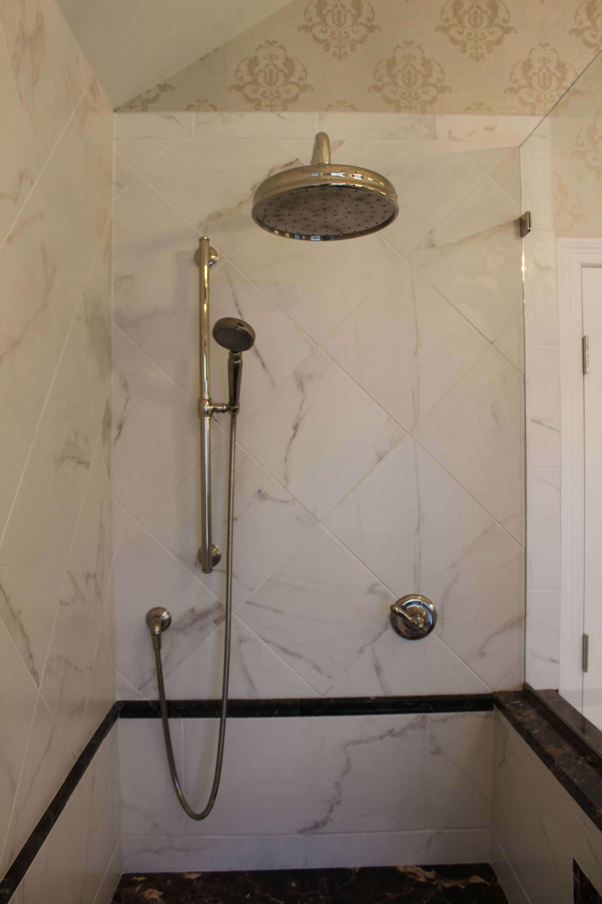 Bathroom shower with marble-look tile, chrome rain showerhead, handheld shower, and grab bar.