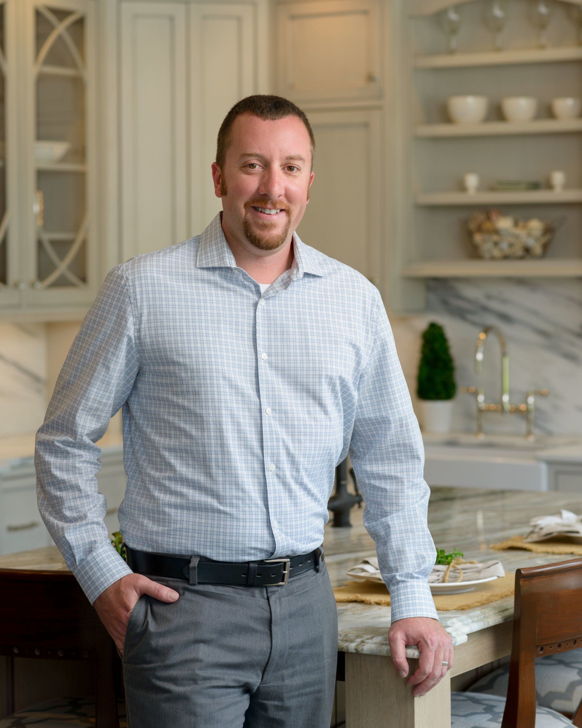 Man standing in a bright kitchen, wearing a light blue shirt and gray pants, smiling beside a counter.