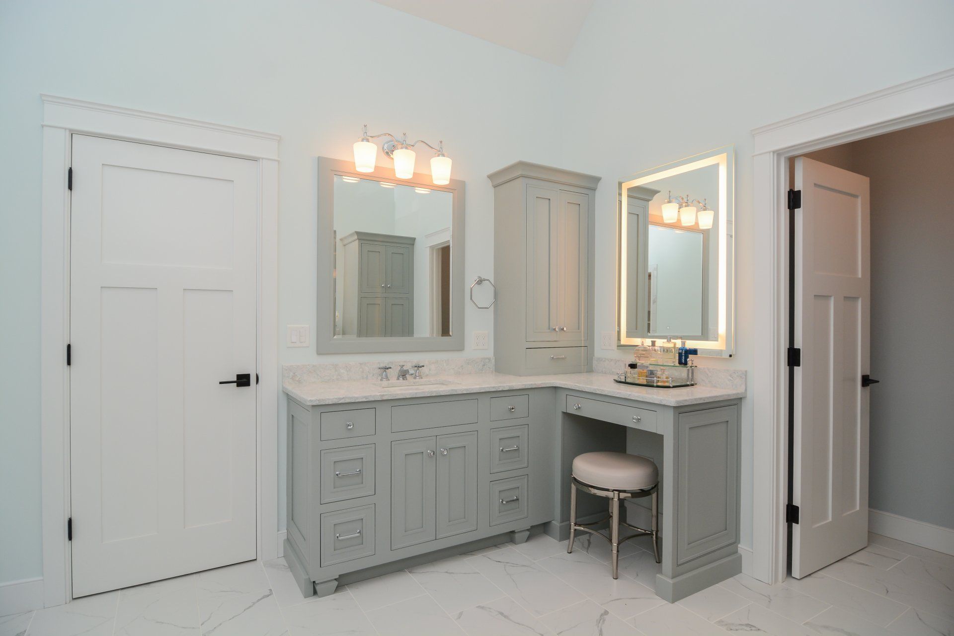 Bright white bathroom vanity with double mirrors, lights, and a small stool beside a closed door.
