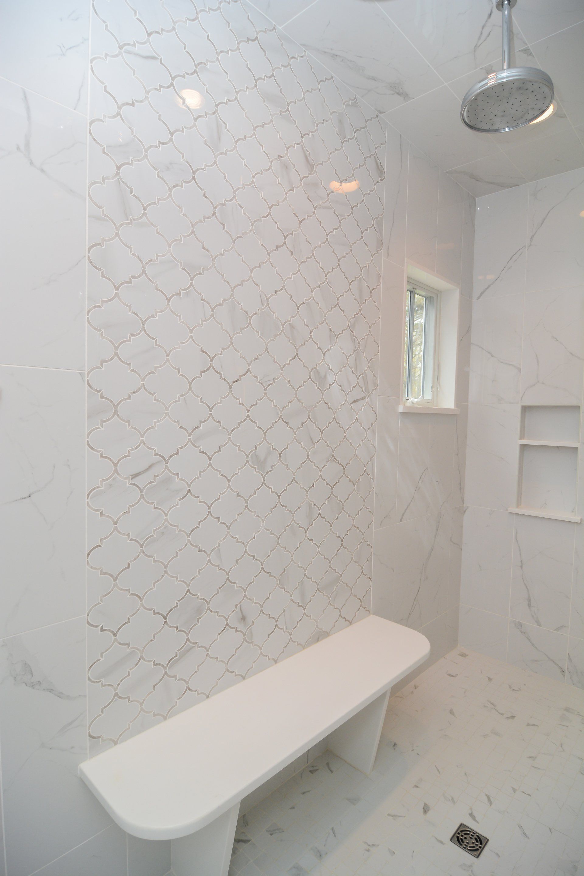 Modern white-tiled bathroom with a bench, showerhead, window, and built-in shelf.