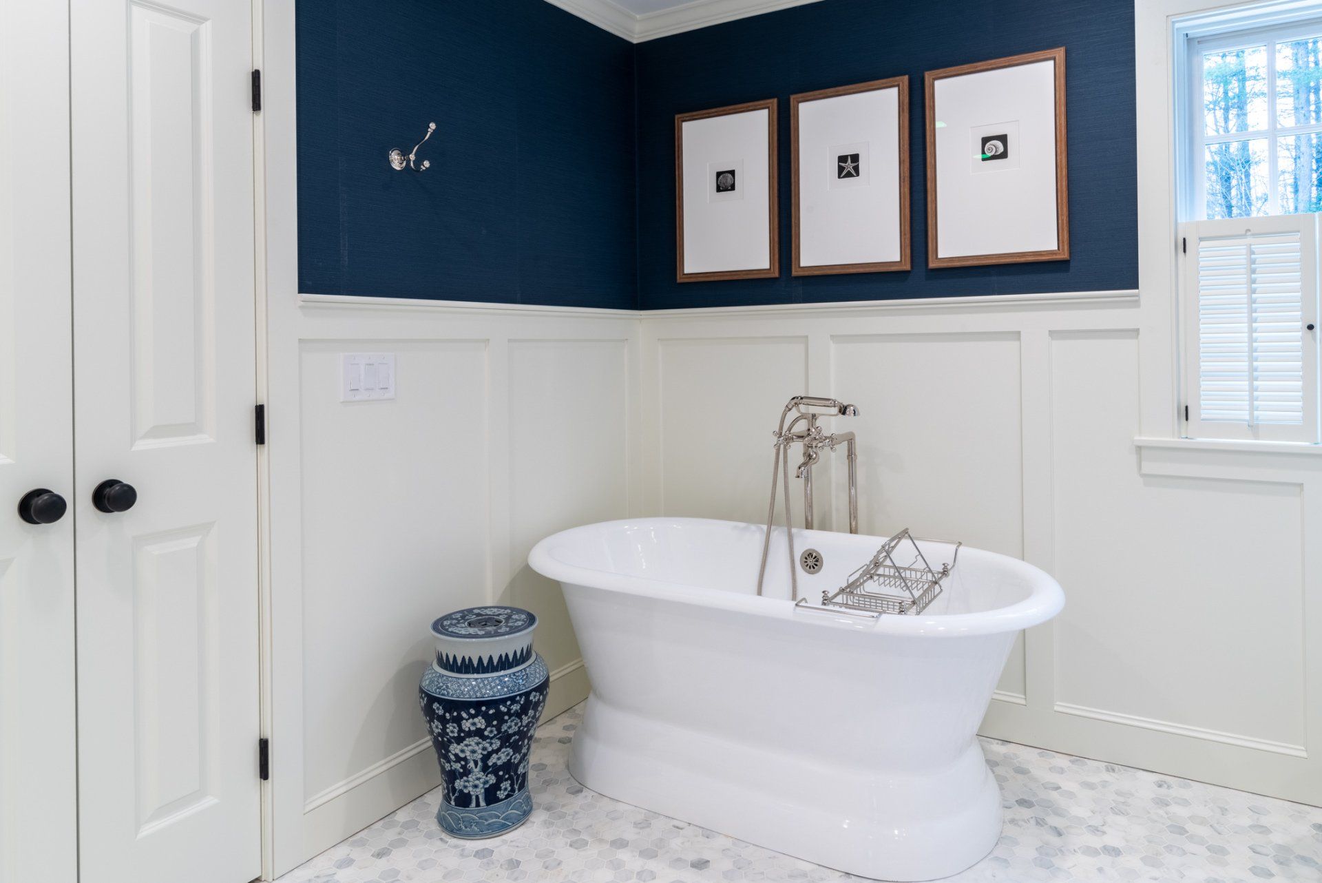 Bright bathroom with a freestanding tub, navy accent wall, white wainscoting, and framed art above the tub.