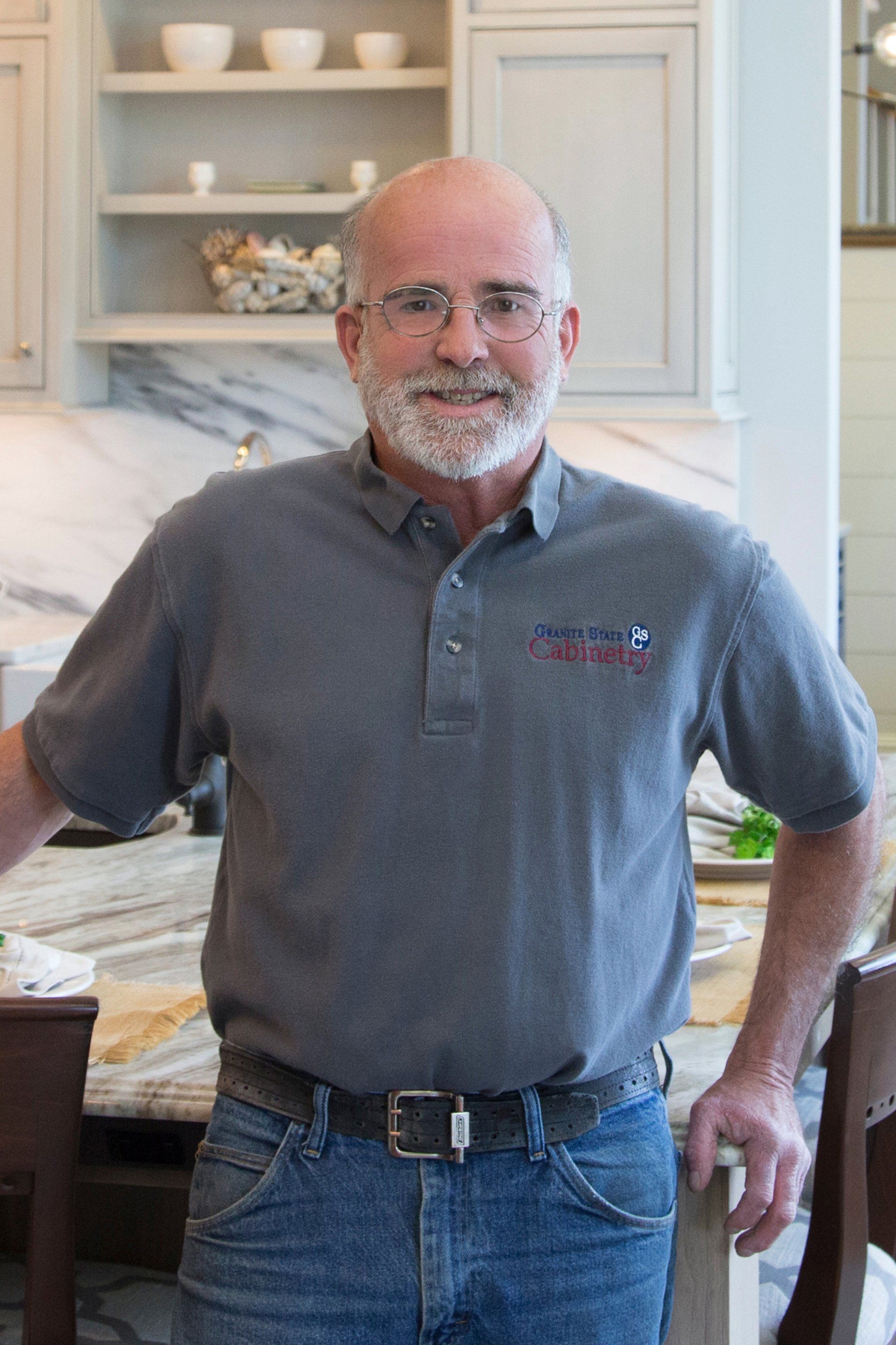 Man standing in a kitchen, wearing a gray polo shirt and jeans, with hands on hips.